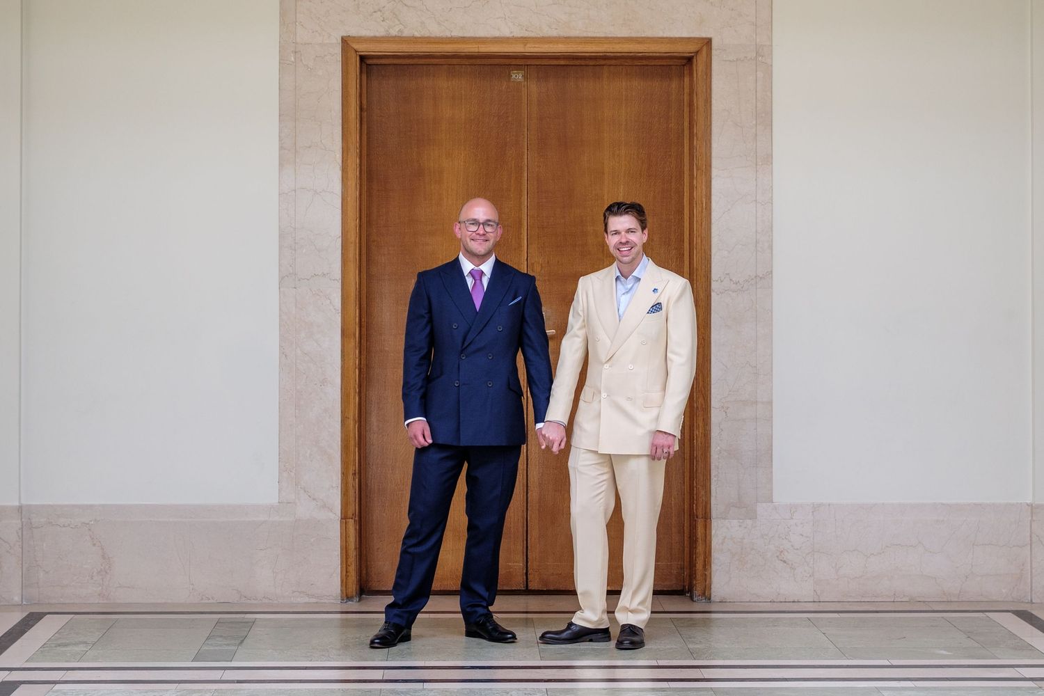 Two people stand in an elegant hallway with arched doorways wearing contrasting dark and light formal attire.