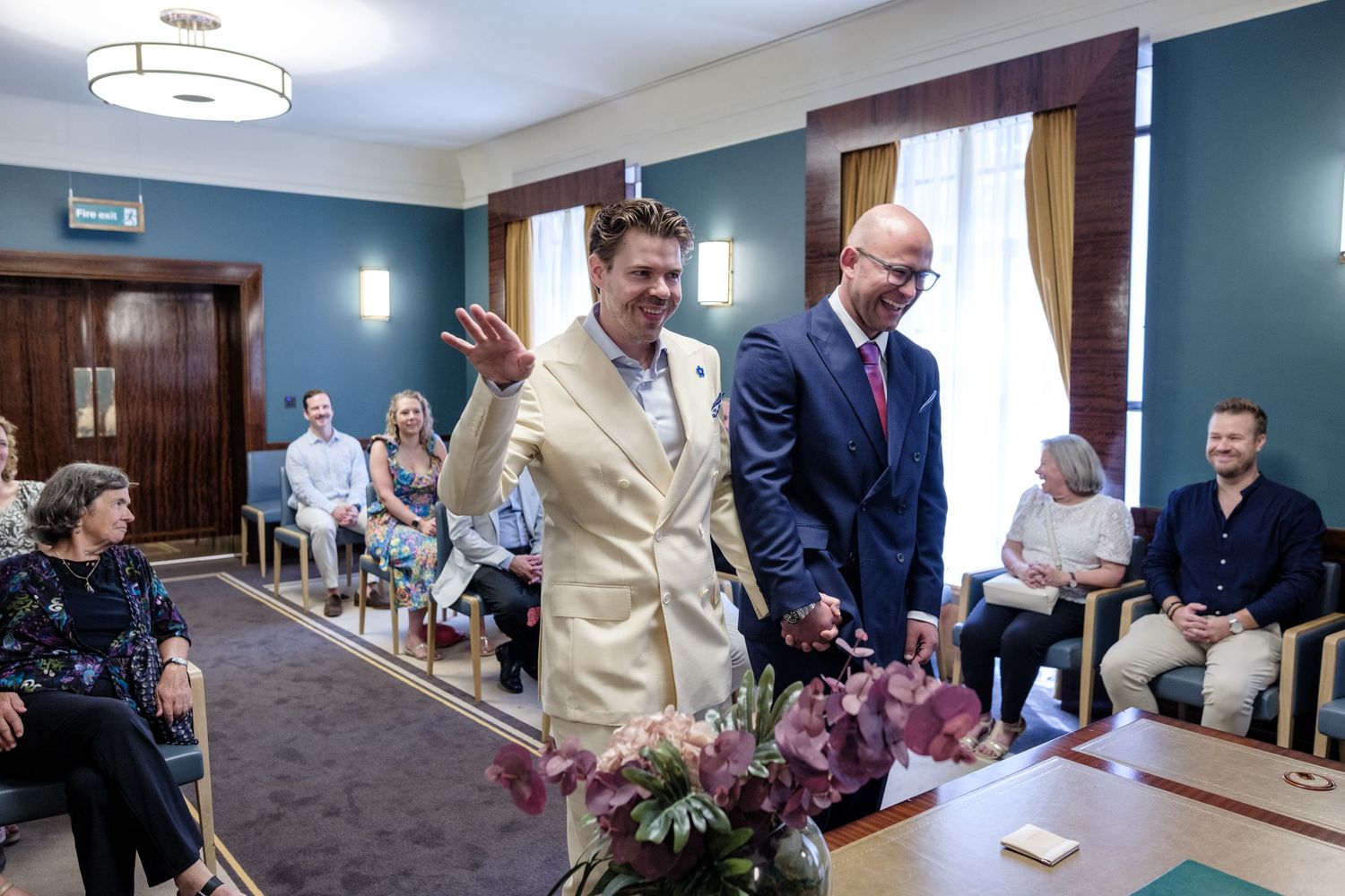 Two people stand together during a wedding ceremony in a room with teal walls and seated guests.