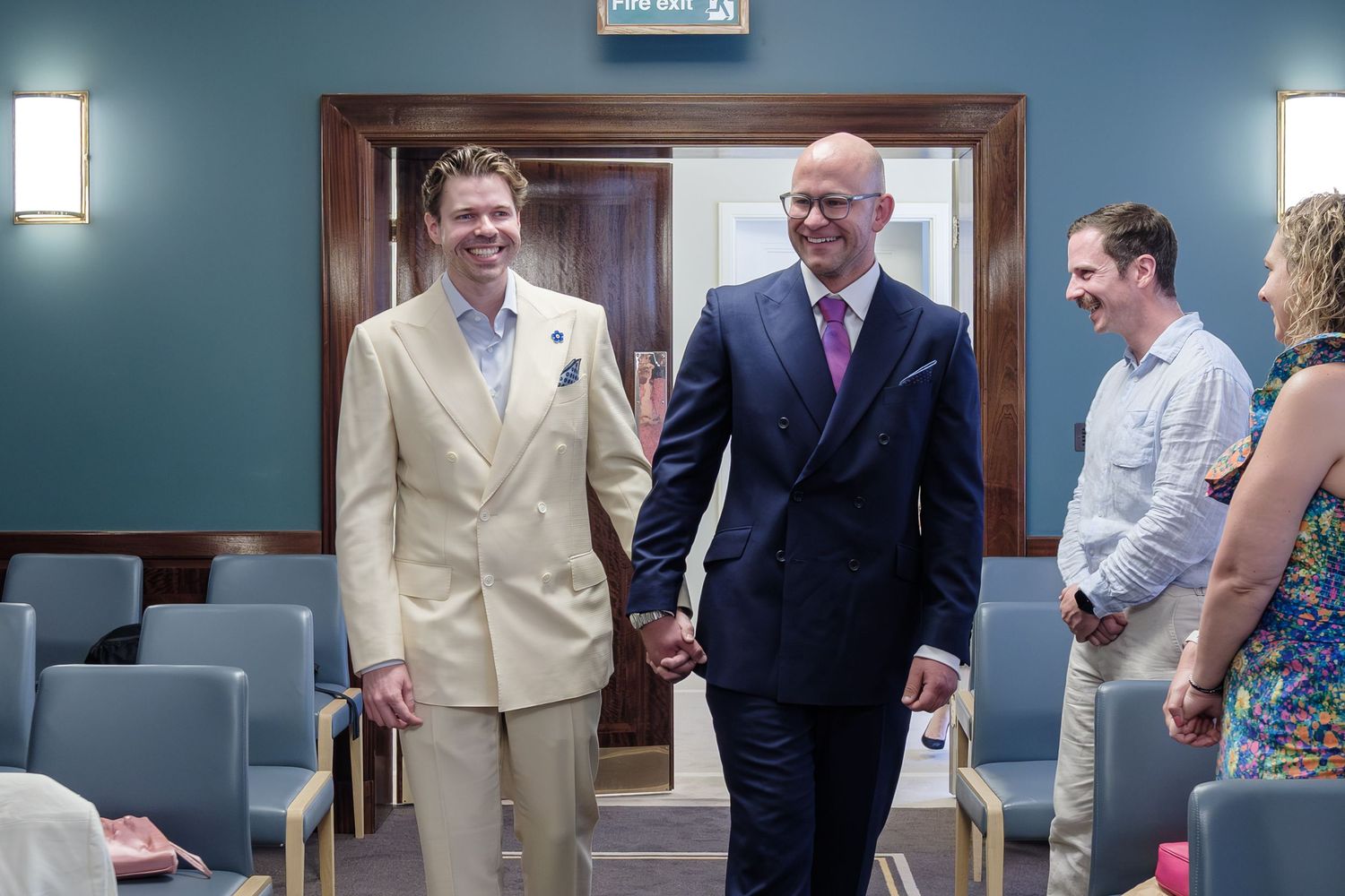 Two people in formal suits, one beige and one navy blue, walking together with smiles in a hallway setting.