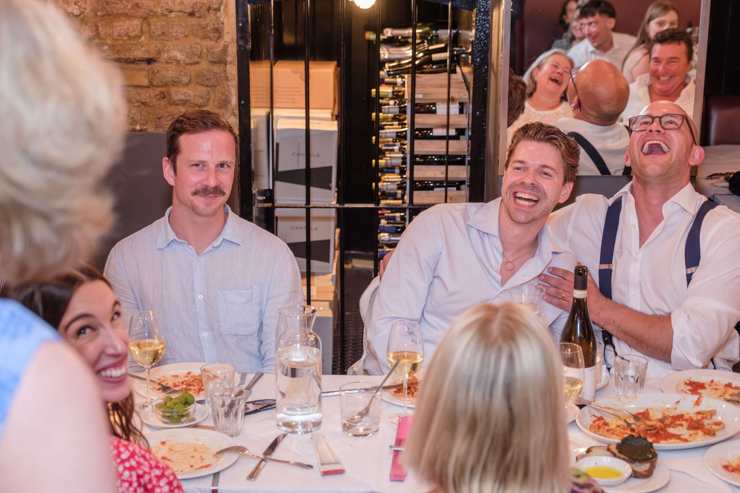 Joyful dinner party guests laughing heartily while enjoying a meal together at a rustic restaurant with brick walls.