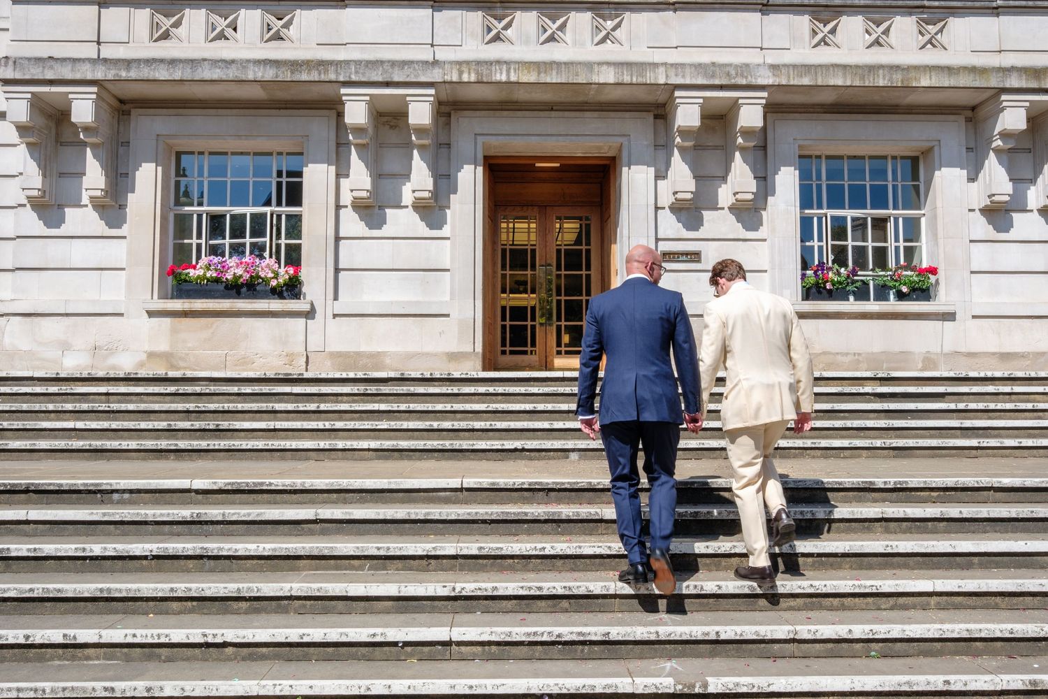 Two people climb stone steps leading to a grand building entrance with ornate architectural details and windows.