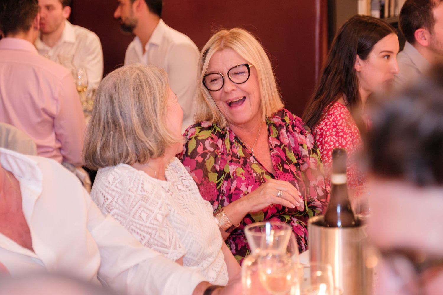 Two people laugh and engage in animated conversation at a social gathering with pink and red tones in the background.