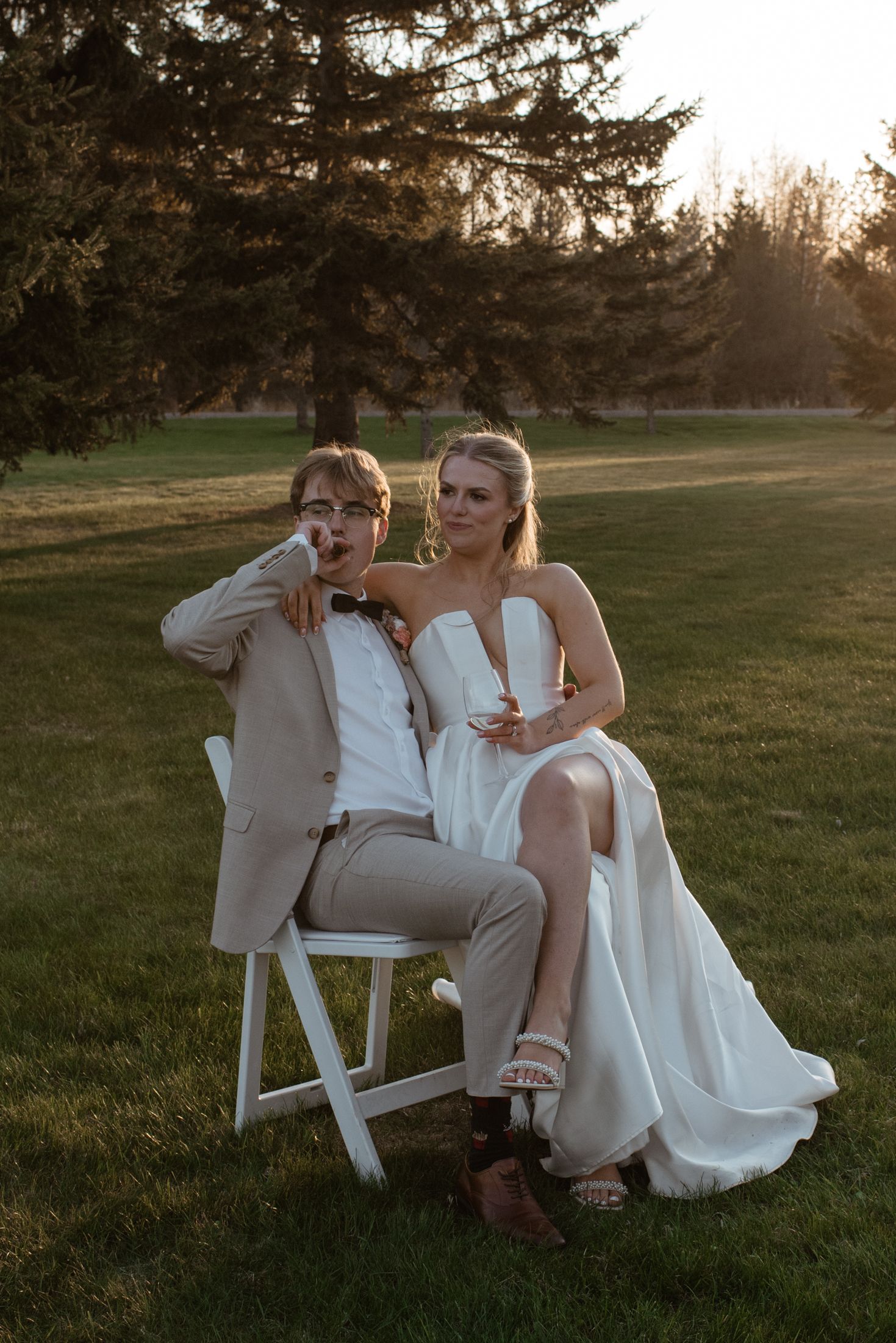 A wedding couple poses together on a white folding chair at sunset in a grassy field with evergreen trees in the background.