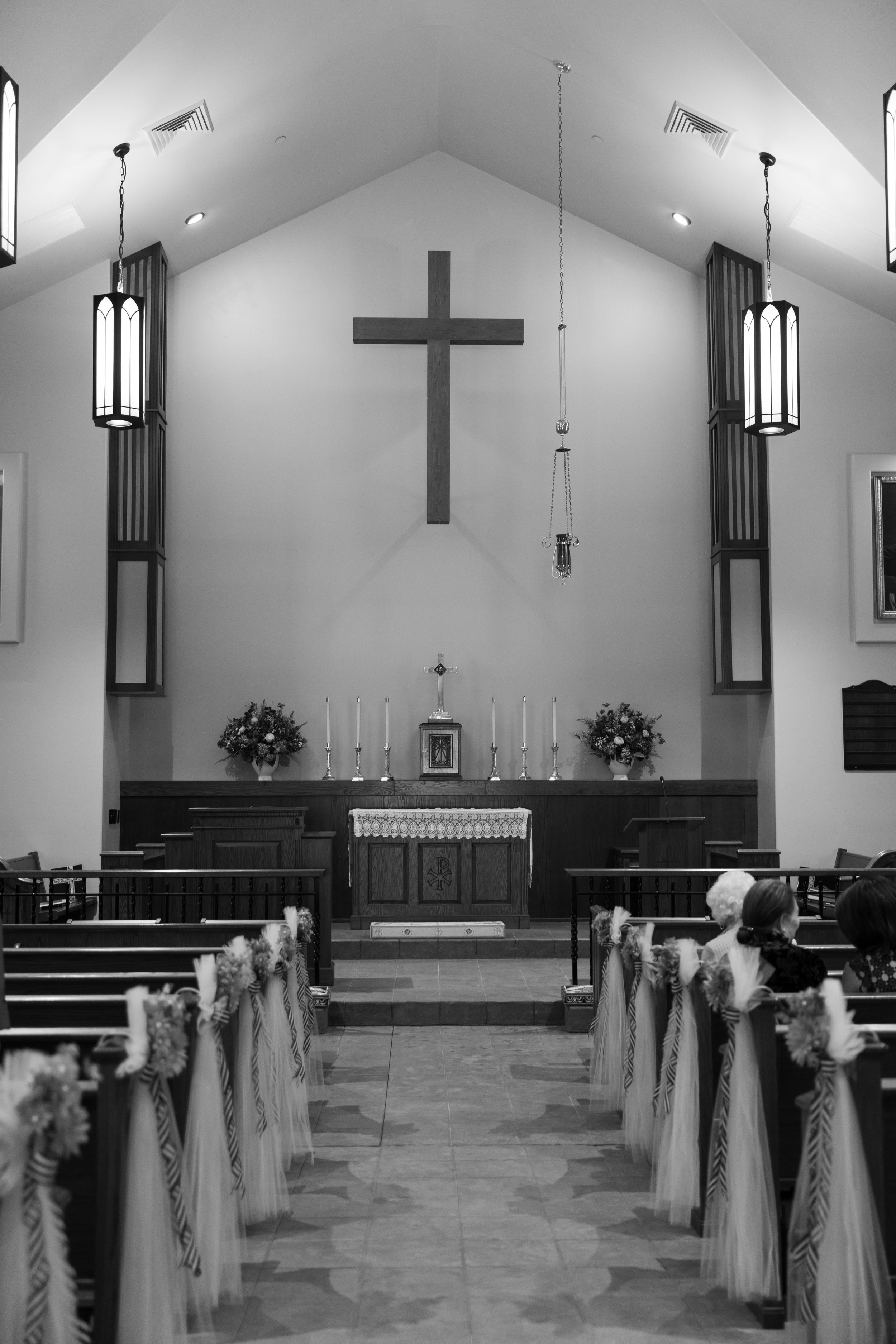 Black and white interior of a church chapel with cross, pews decorated with white ribbons, and floral arrangements at the altar.