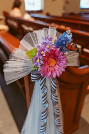 Colorful floral pew decoration with pink gerbera daisy, purple flowers, and white tulle attached with zebra-striped ribbon.