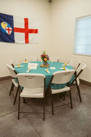 A round table with turquoise tablecloth and folding chairs sits beneath an Episcopal flag in a church meeting room.