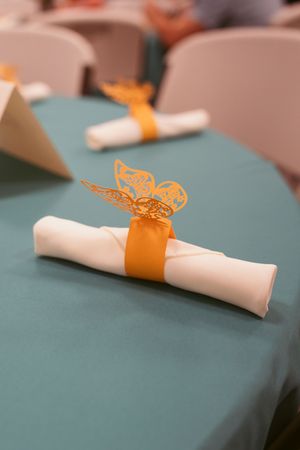 Rolled white napkin tied with orange ribbon and delicate butterfly decoration on teal tablecloth at formal event.