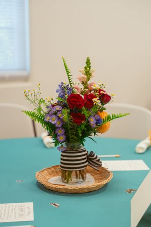 A colorful floral centerpiece with red roses and blue flowers in a striped mason jar sits on a woven placemat against turquoise.