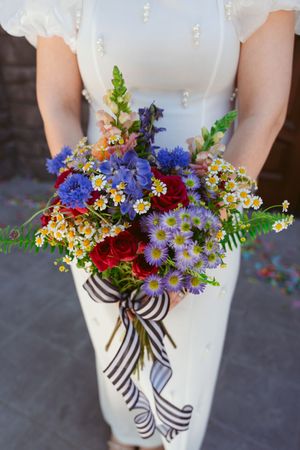 A vibrant wildflower bouquet with blue cornflowers, red roses and yellow blooms tied with a striped ribbon against a white dress.