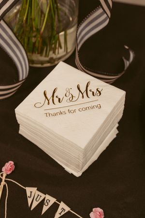 Stack of white wedding napkins with 'Mr & Mrs - Thanks for coming' text in gold lettering and decorative ribbon nearby.