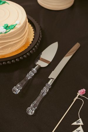 Close-up view of silver cake cutting utensils beside a frosted white cake with green garnish on a dark background.
