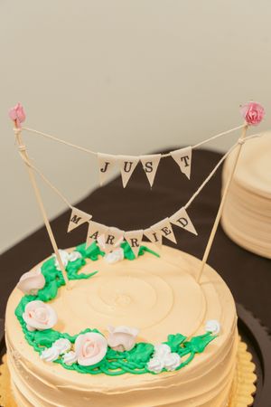 Wedding cake topped with a 'JUST MARRIED' banner and decorated with green leaves and white roses around the edge.