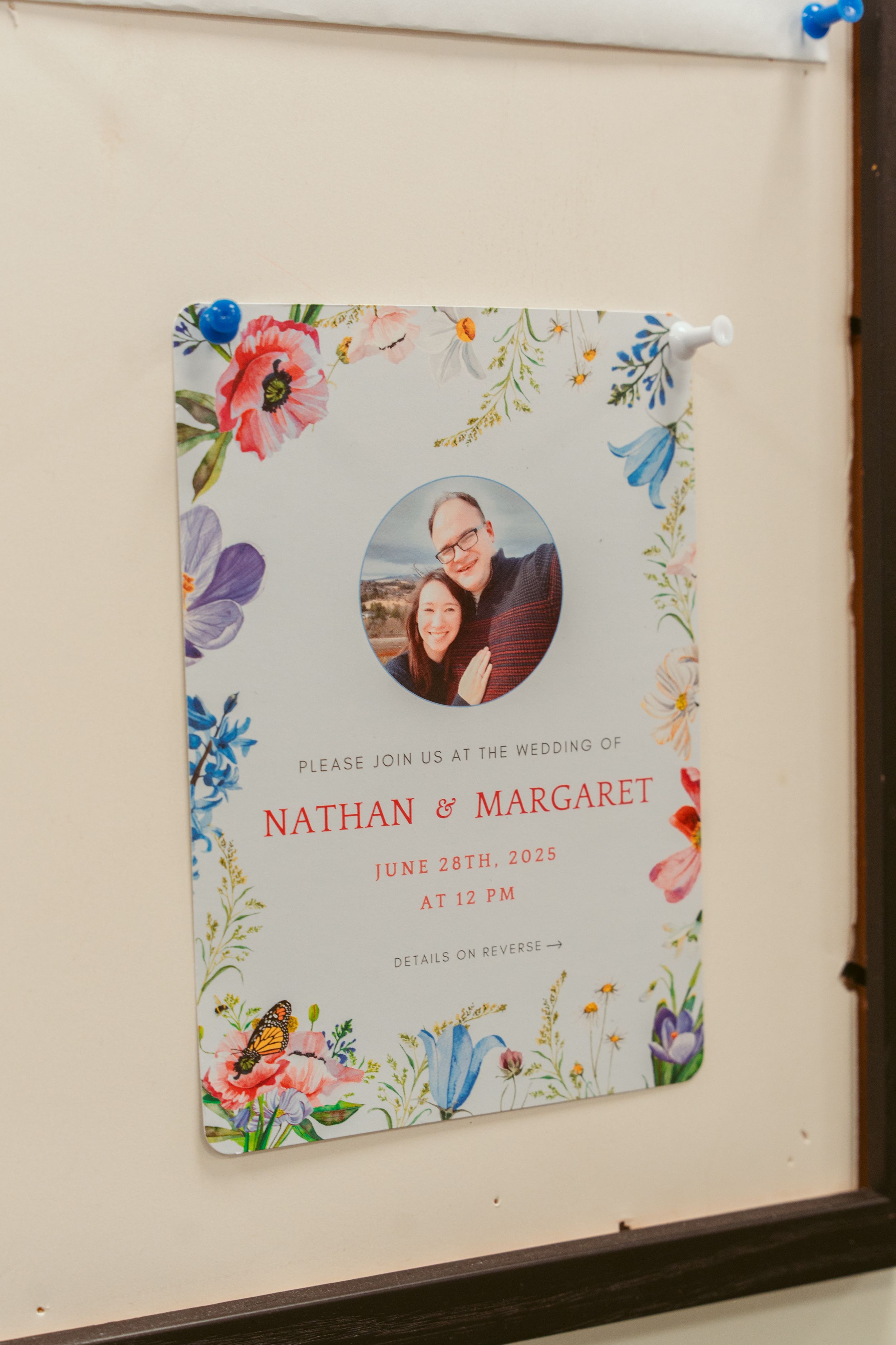 A floral-bordered wedding invitation with a circular photo in the center features delicate watercolor blooms in pink and blue.
