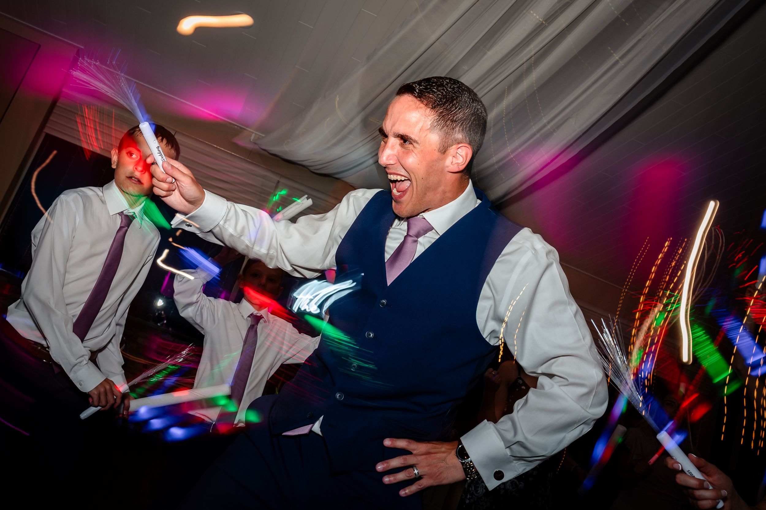 Wedding reception dance floor illuminated with purple lighting and guests dancing with glow sticks.