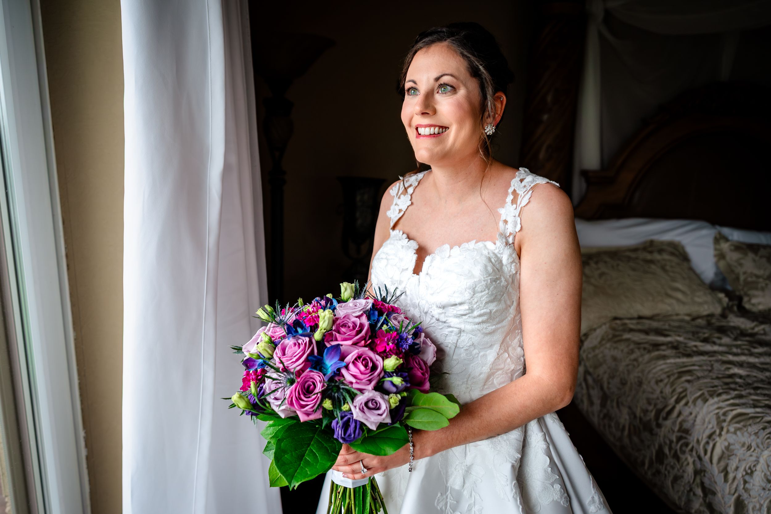 Bride holding vibrant purple and pink floral bouquet by window.