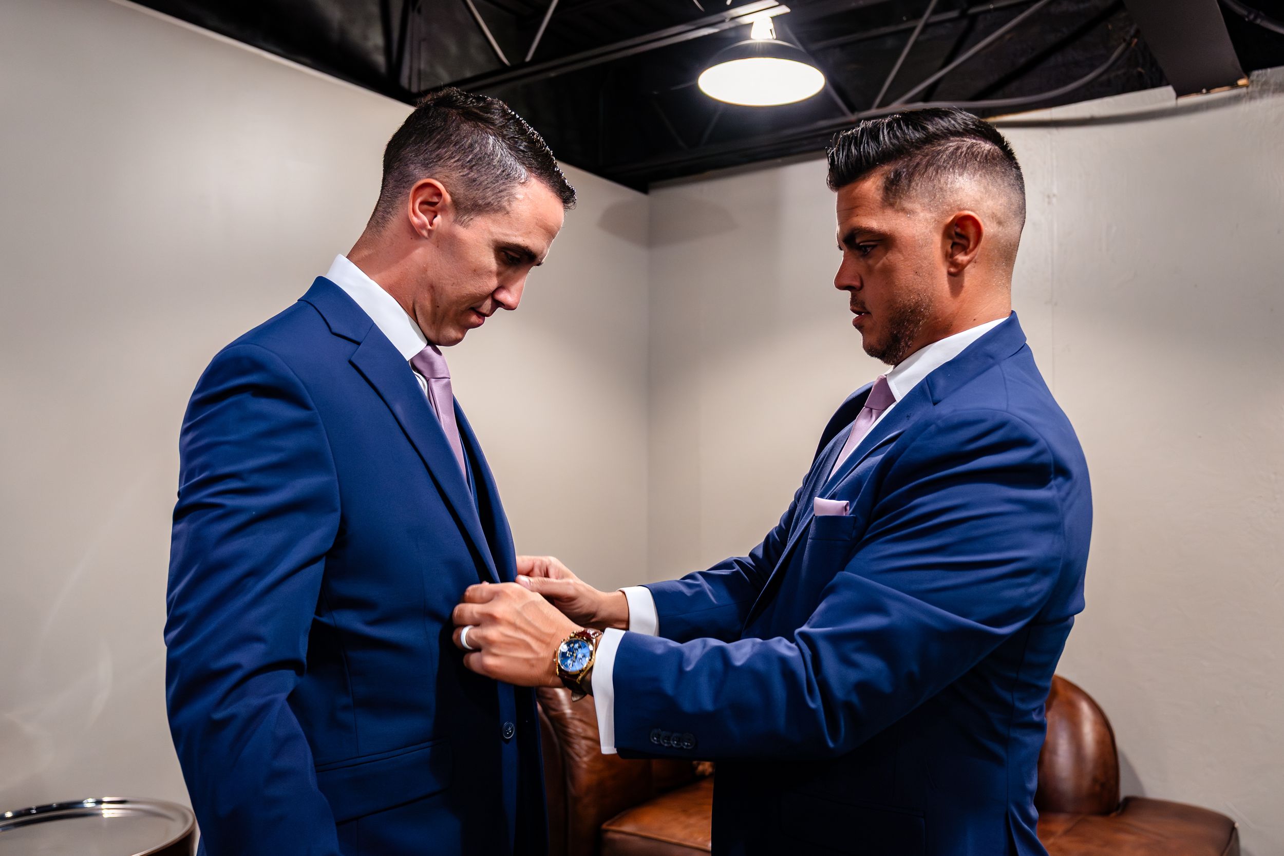 Two people in navy suits adjusting their formal attire in preparation room.