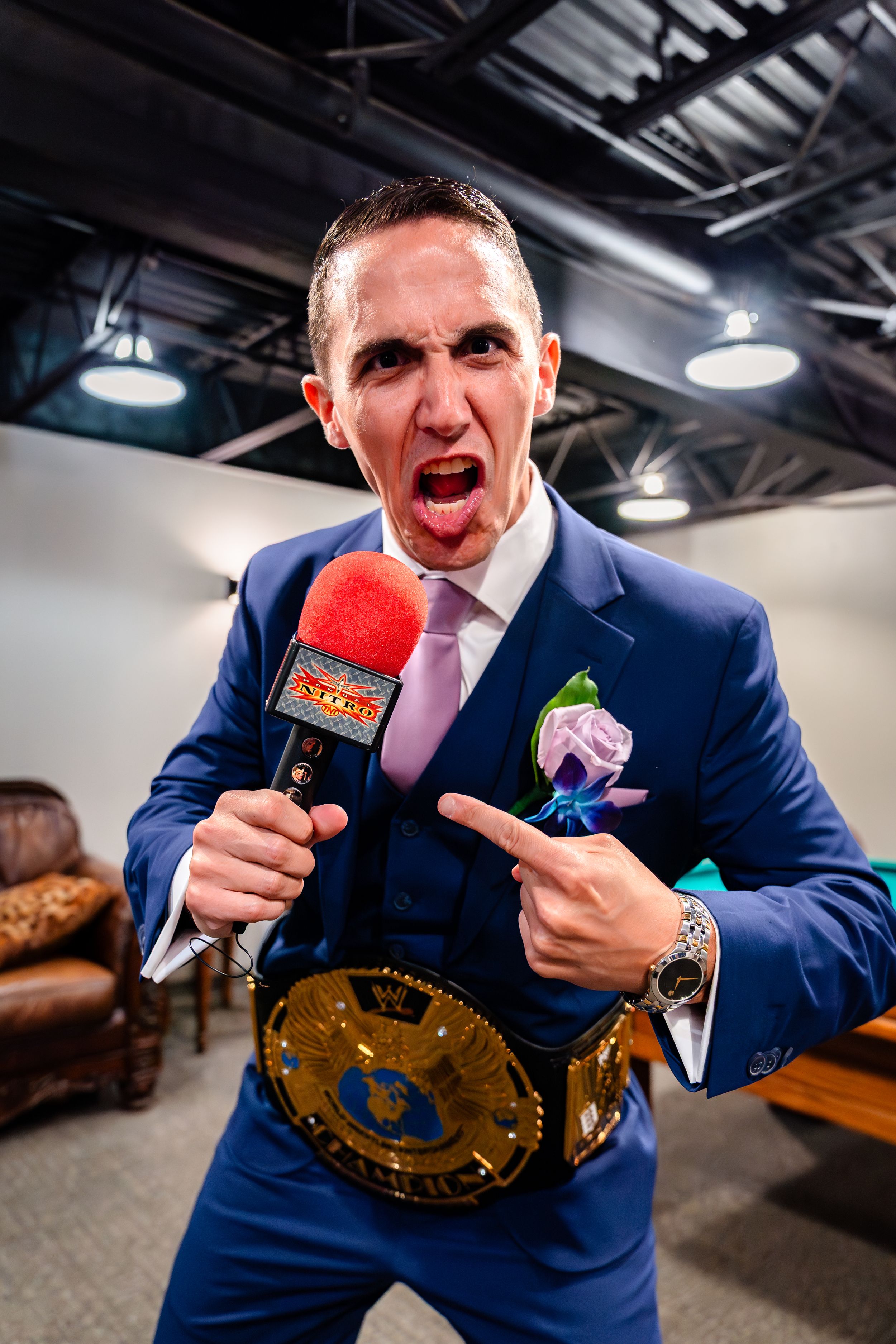 Person in blue suit poses with championship belt and colorful microphones in indoor setting.