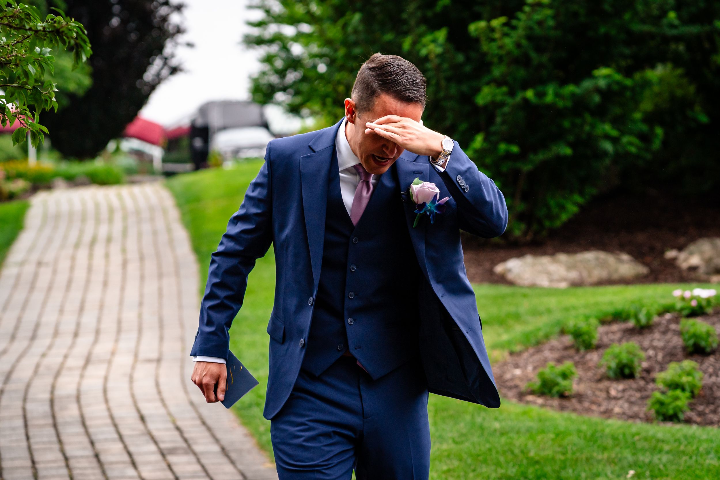 Groom in navy suit walking along garden pathway during wedding photoshoot.