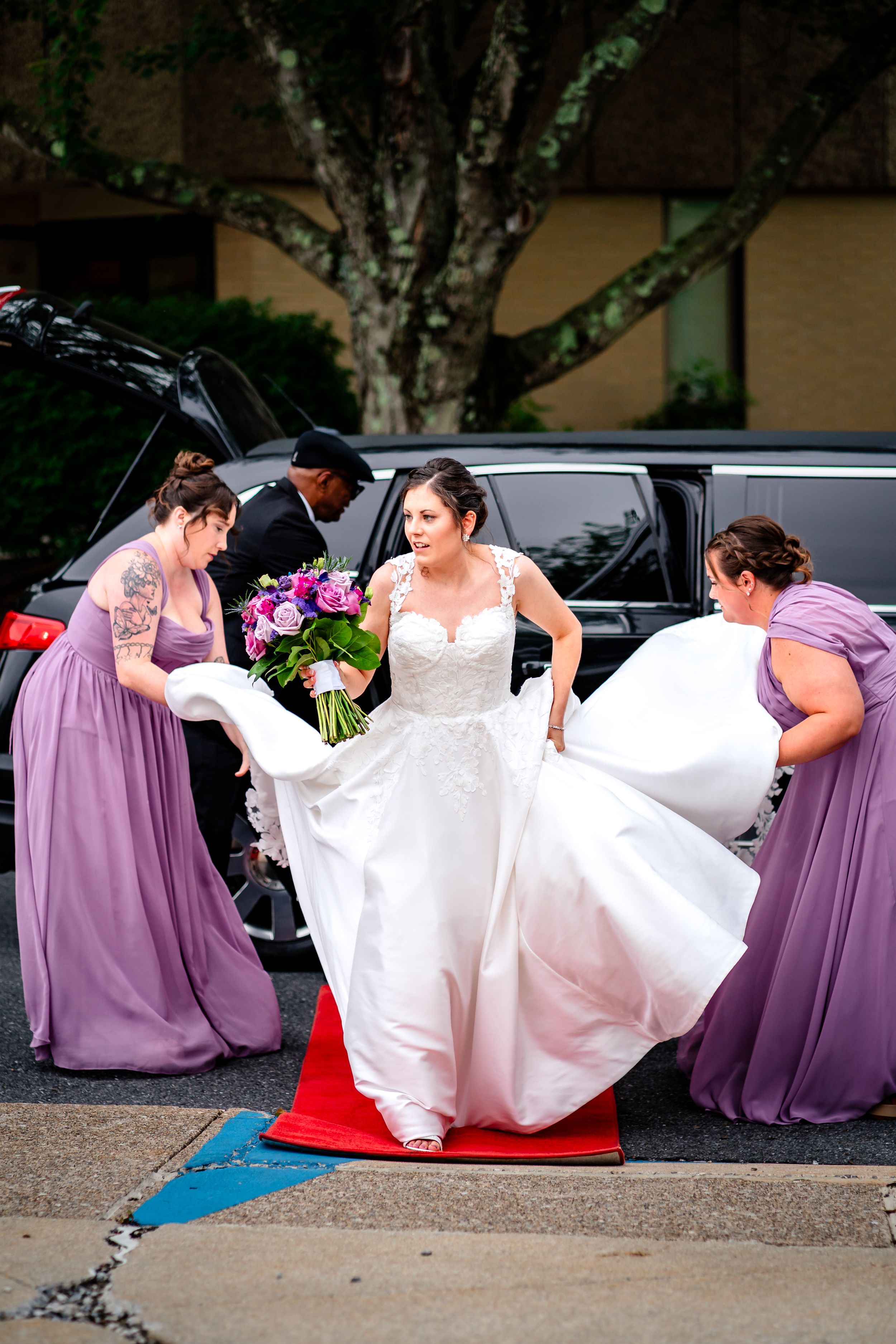Bridesmaids in purple dresses assisting with the bride's wedding gown outdoors.