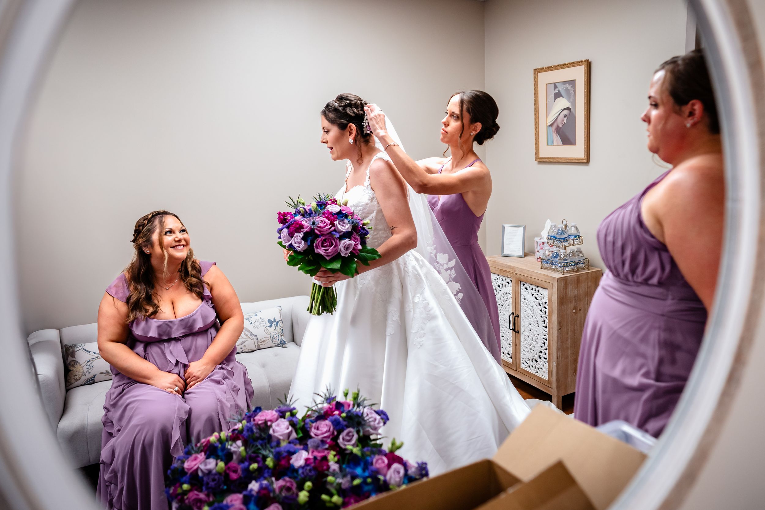 Wedding preparation scene through mirror reflection showing purple bridesmaid dresses and floral bouquets.