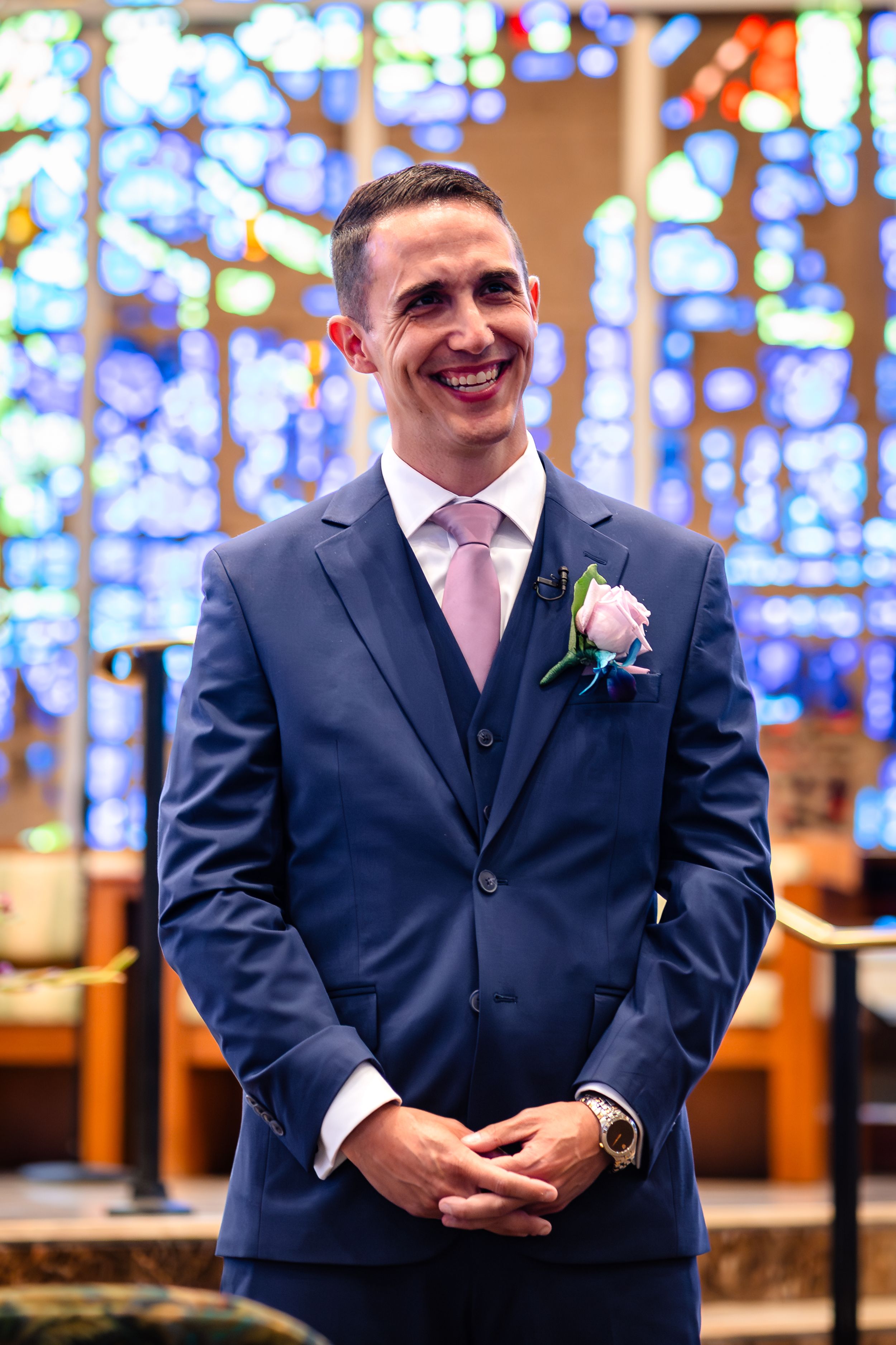Groomsmen in navy suits standing before colorful stained glass windows in church.