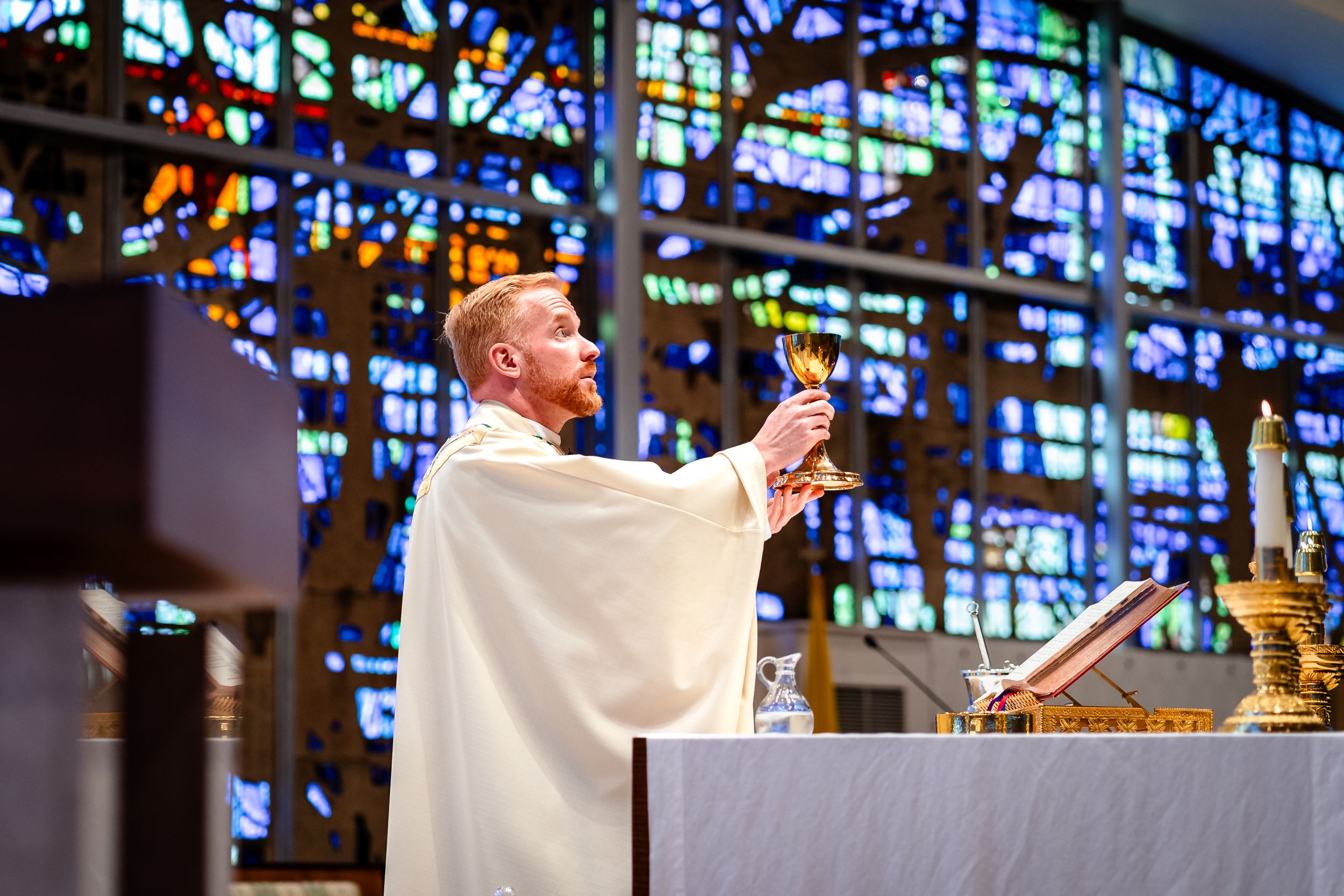 Catholic priest in white robes conducting ceremony at altar with colorful stained glass backdrop.