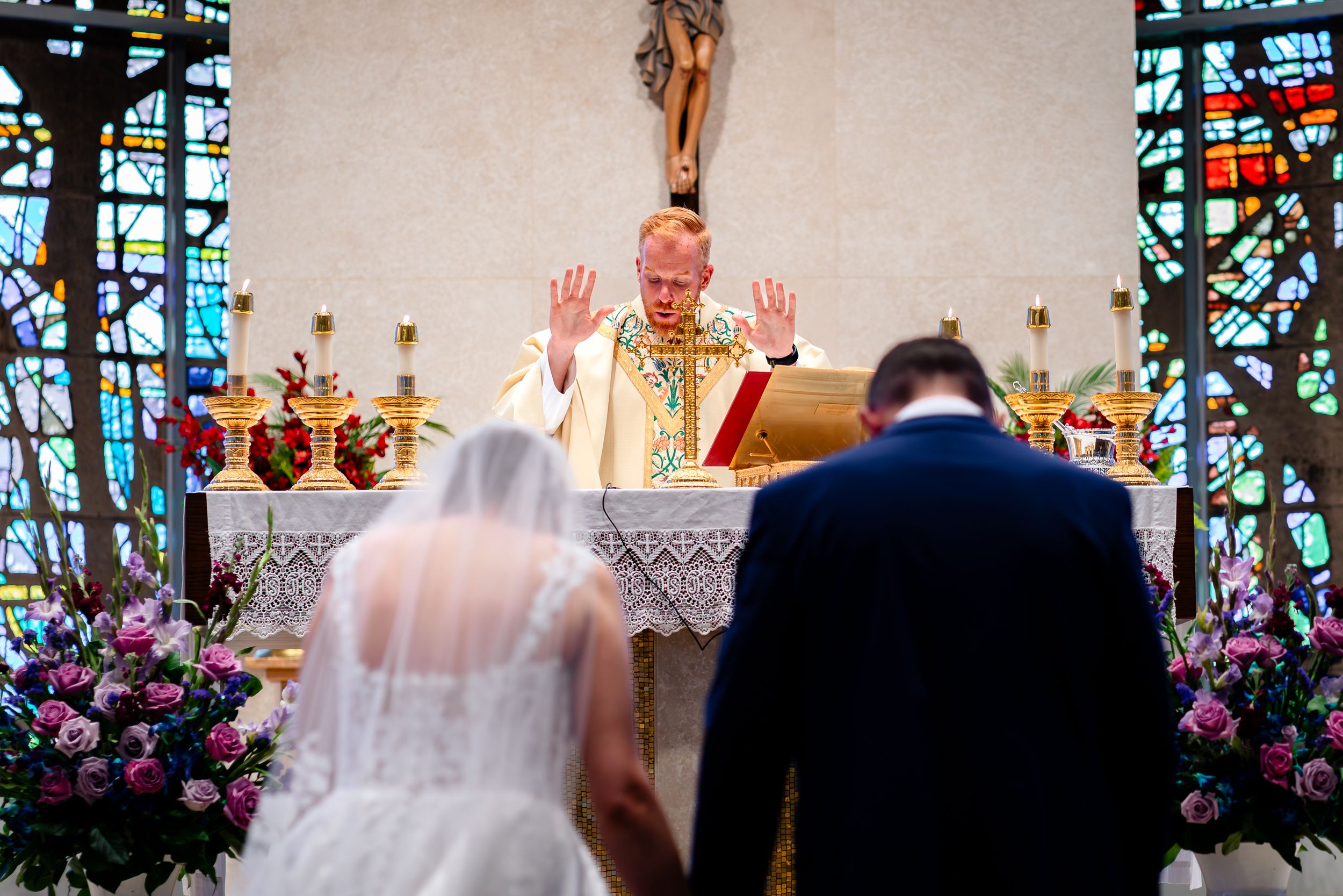 Traditional Catholic wedding blessing at altar surrounded by flowers and stained glass windows.