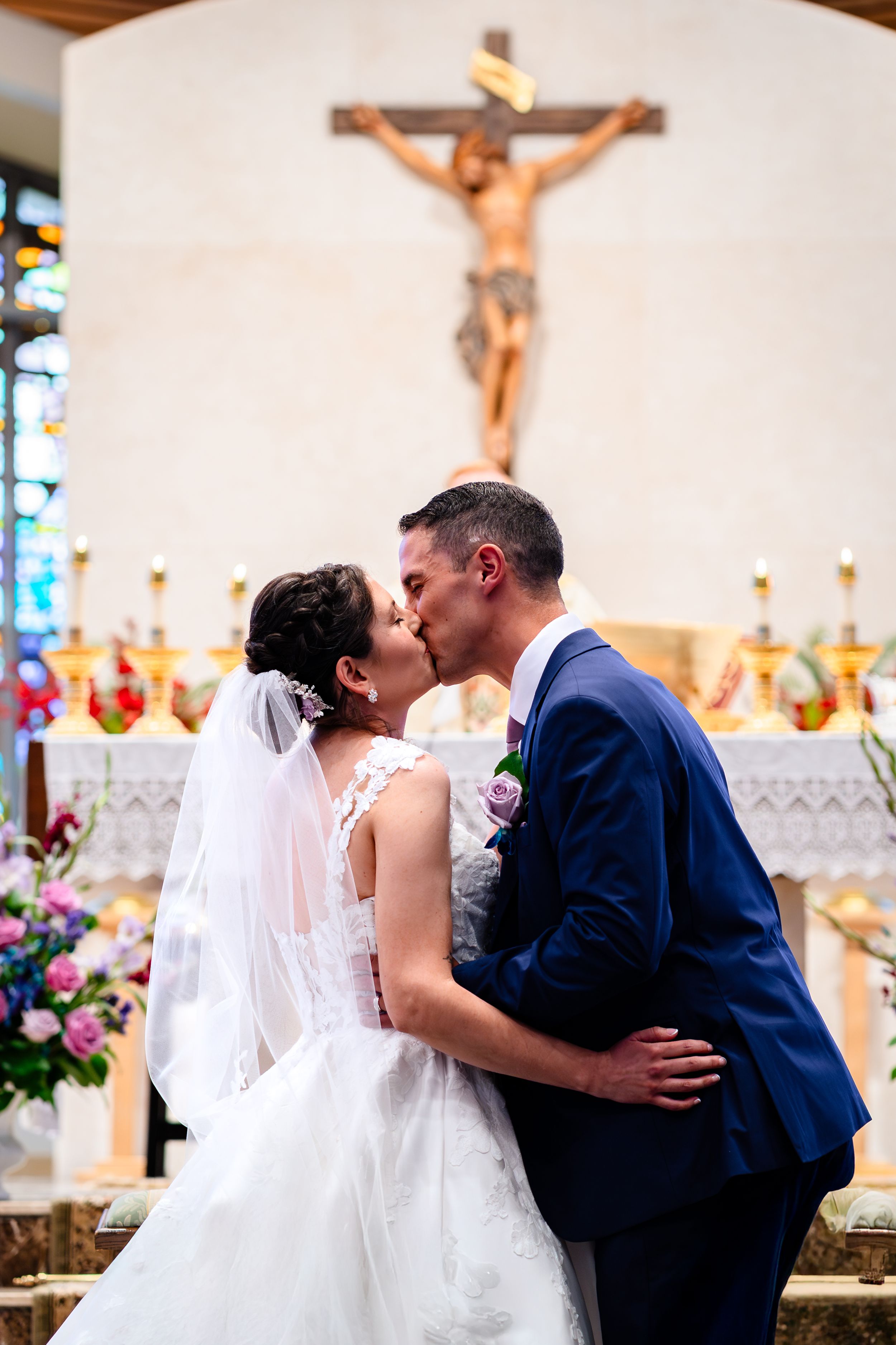 Romantic wedding kiss at church altar beneath crucifix with floral arrangements.