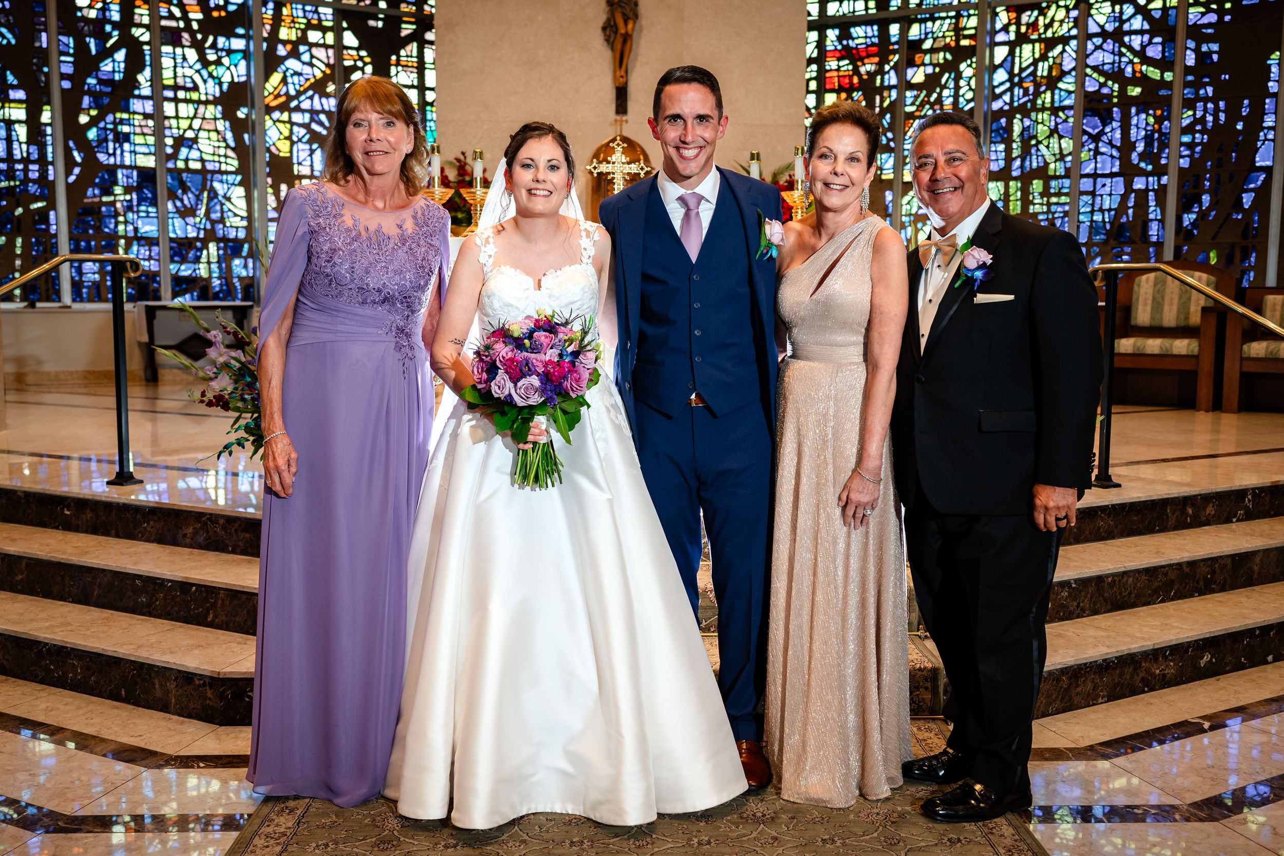 Wedding party in formal attire poses inside church with beautiful stained glass windows in background.