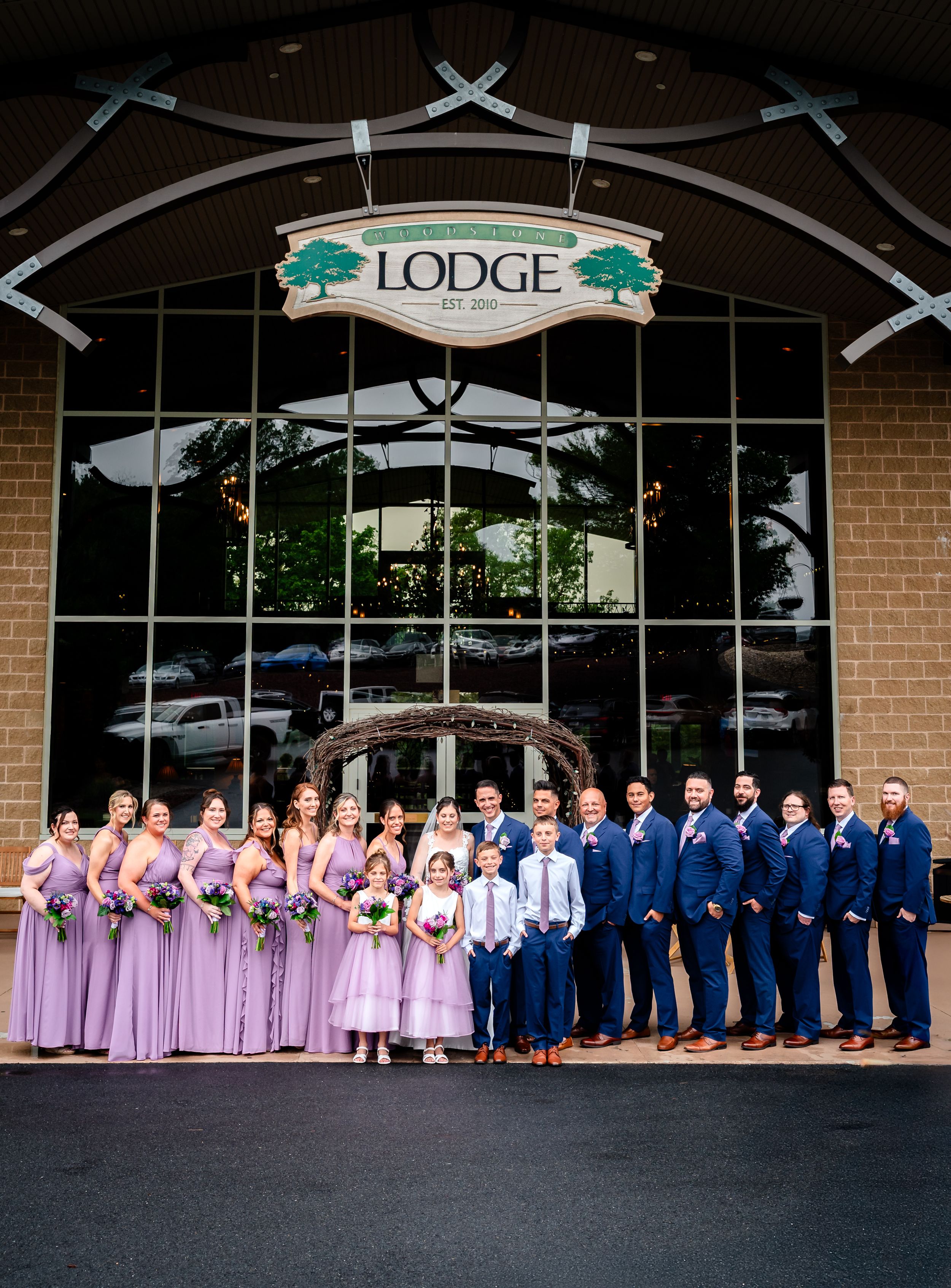 Large wedding party in navy and lavender attire lined up outside The Lodge venue entrance.