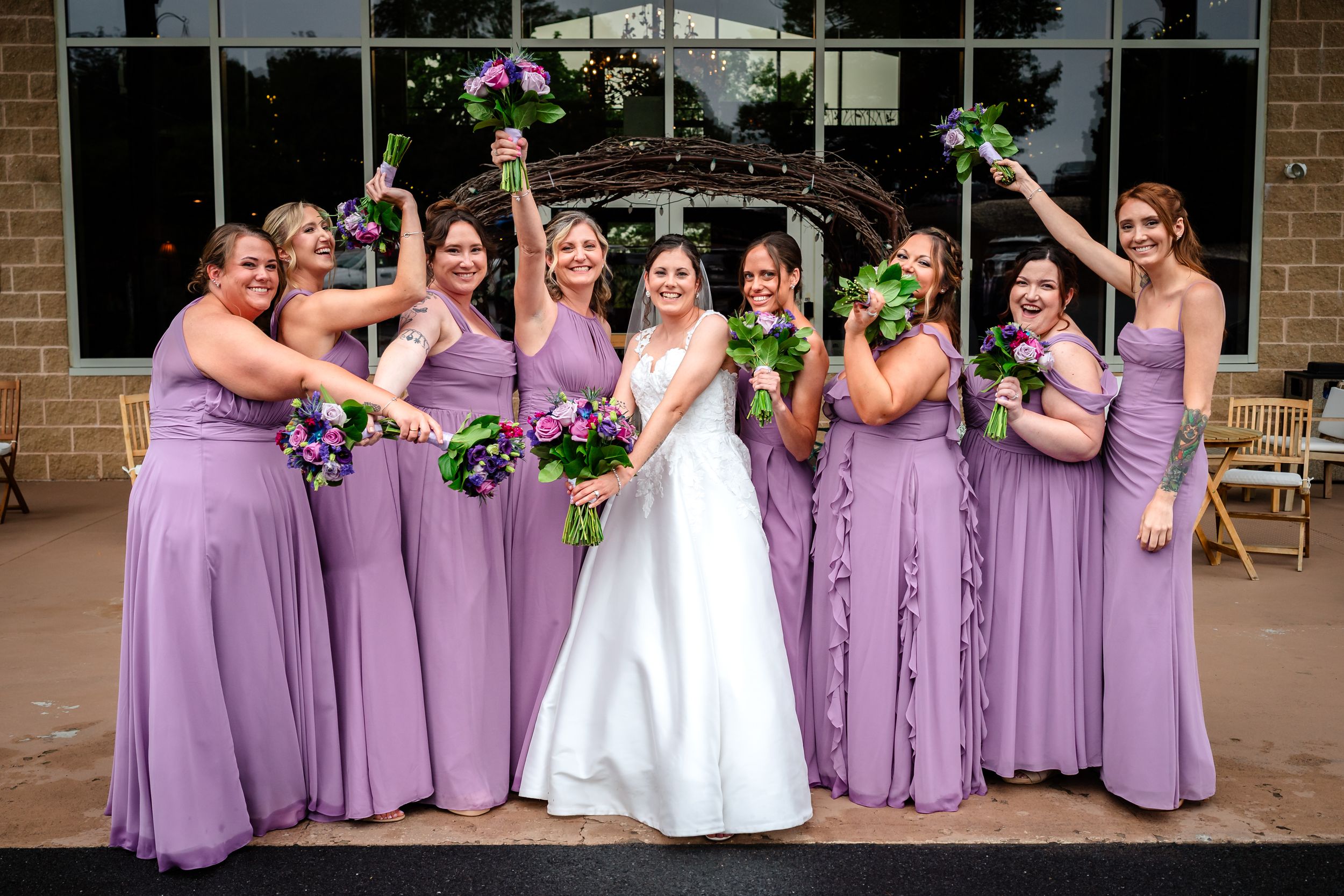 Wedding party in lavender dresses celebrating outside brick venue with large windows.