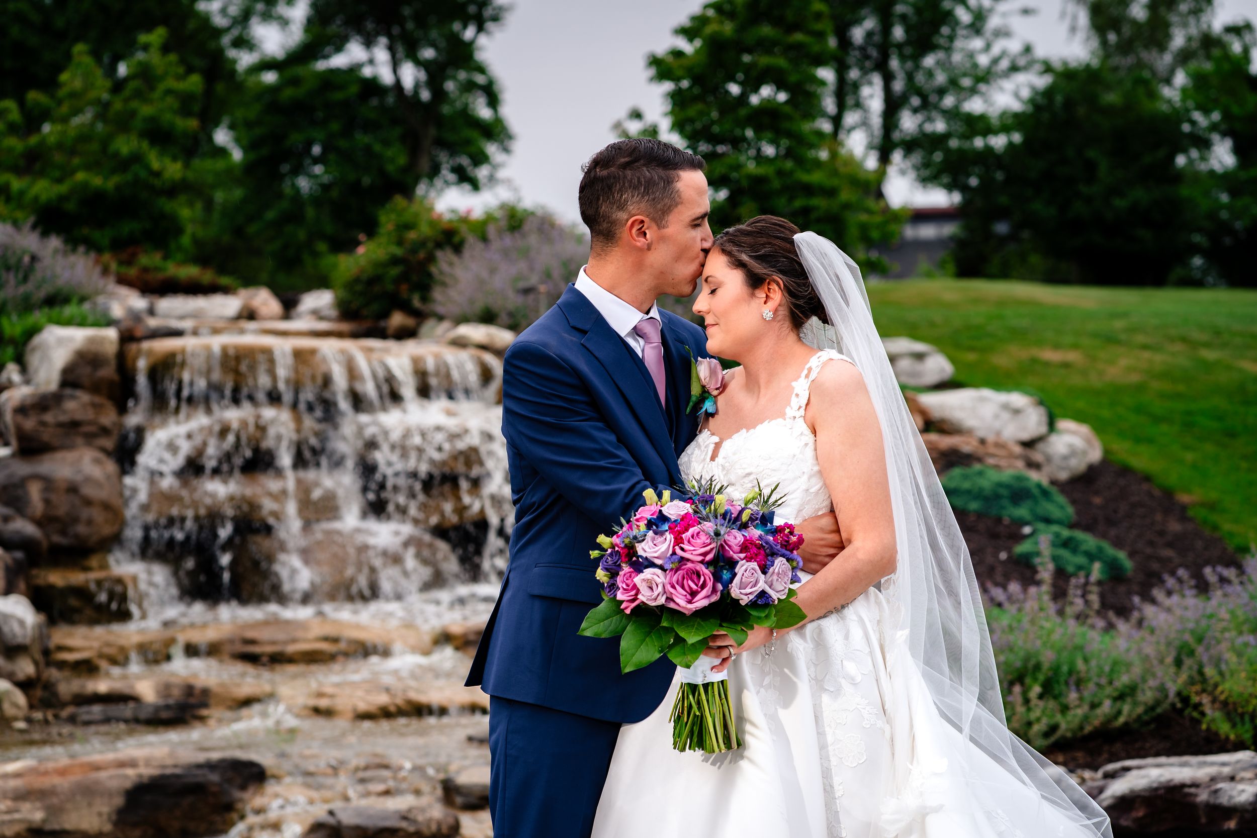 Wedding couple poses by natural stone waterfall feature in landscaped garden setting.