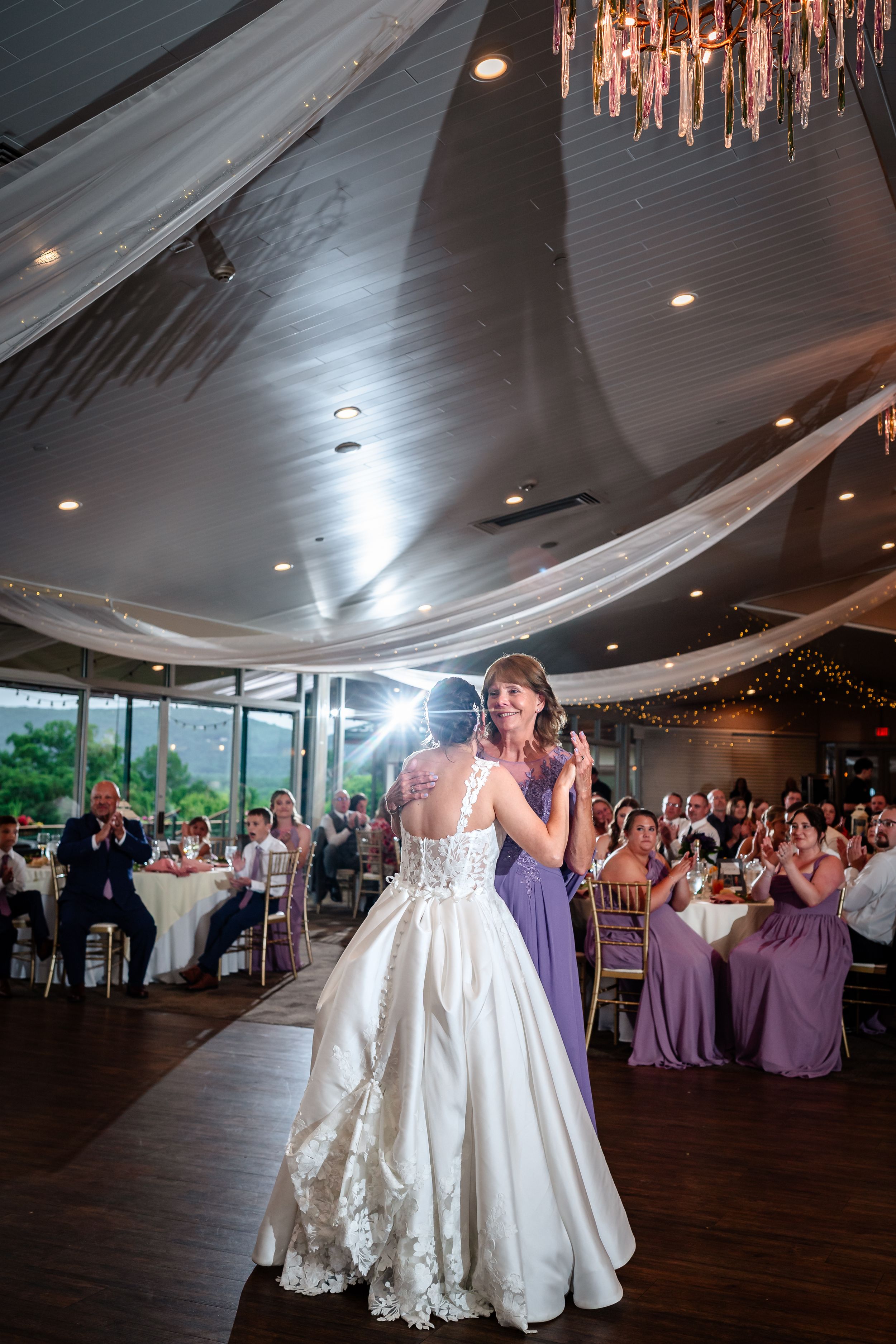 Bride in lace gown stands in spotlight during reception while guests applaud.