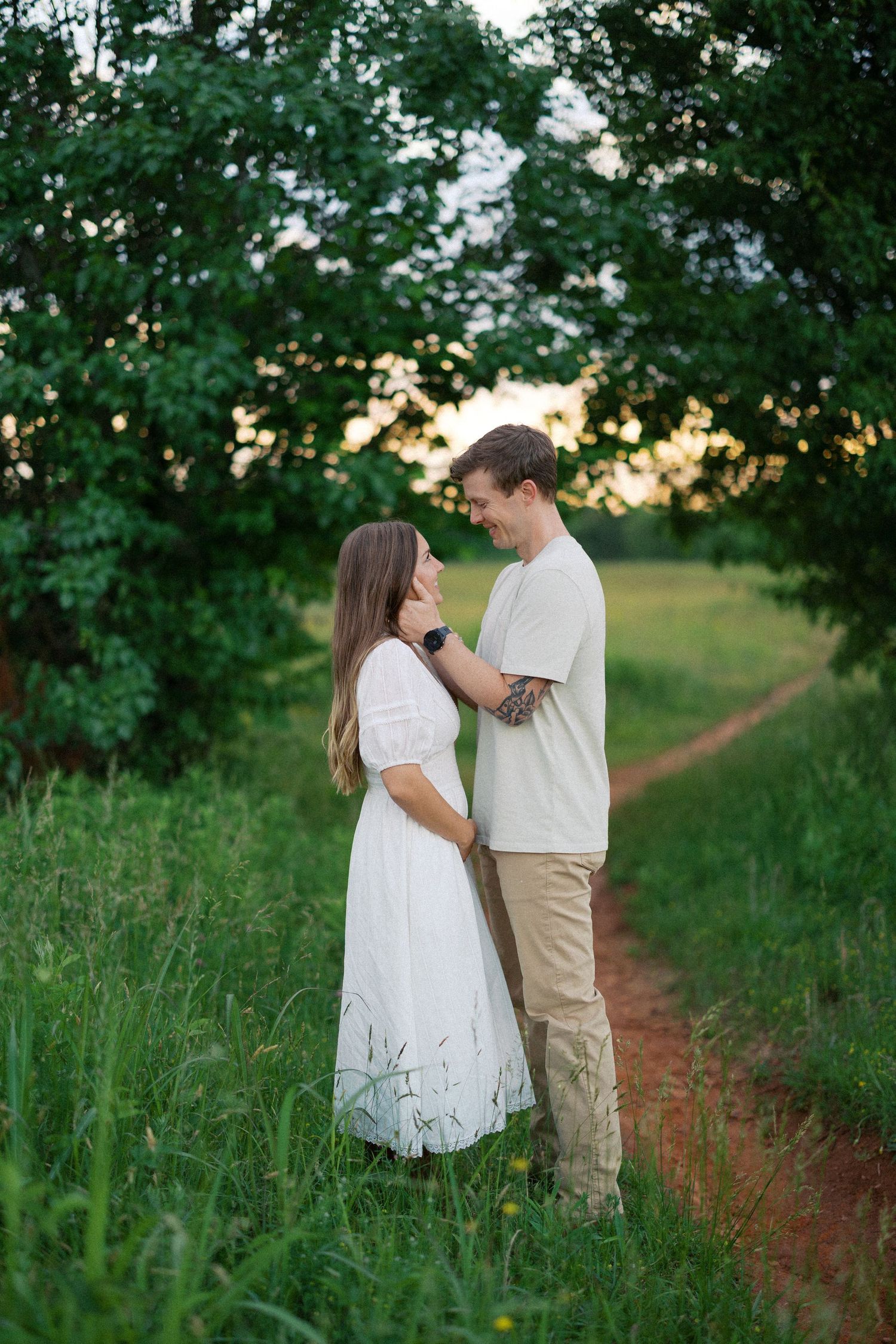 A couple shares a tender moment on a rustic dirt path surrounded by lush green foliage at sunset.