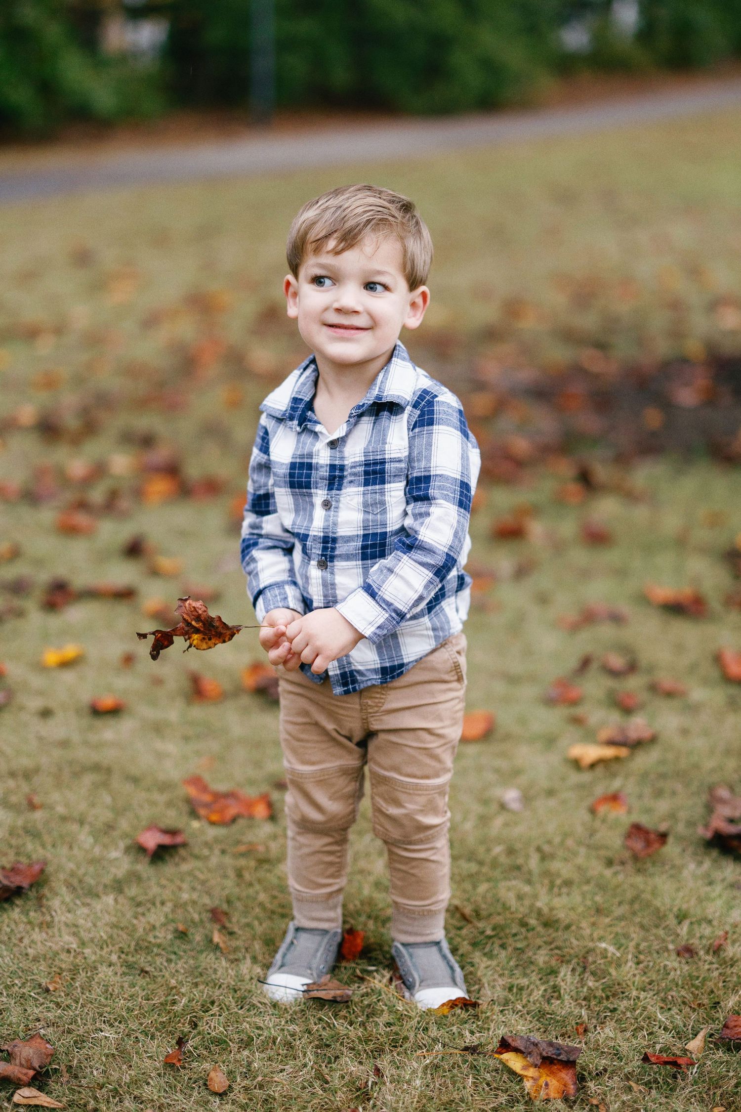 Small child wearing blue plaid shirt and khaki pants stands playfully on grass covered in autumn leaves.