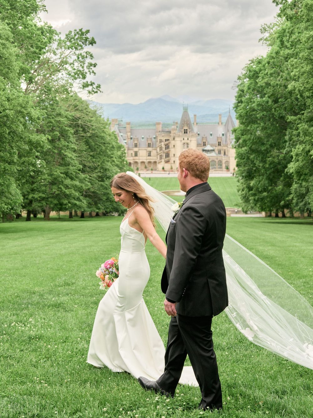 Wedding couple walking across a lush green lawn with their flowing veil trailing behind them towards a grand castle estate.