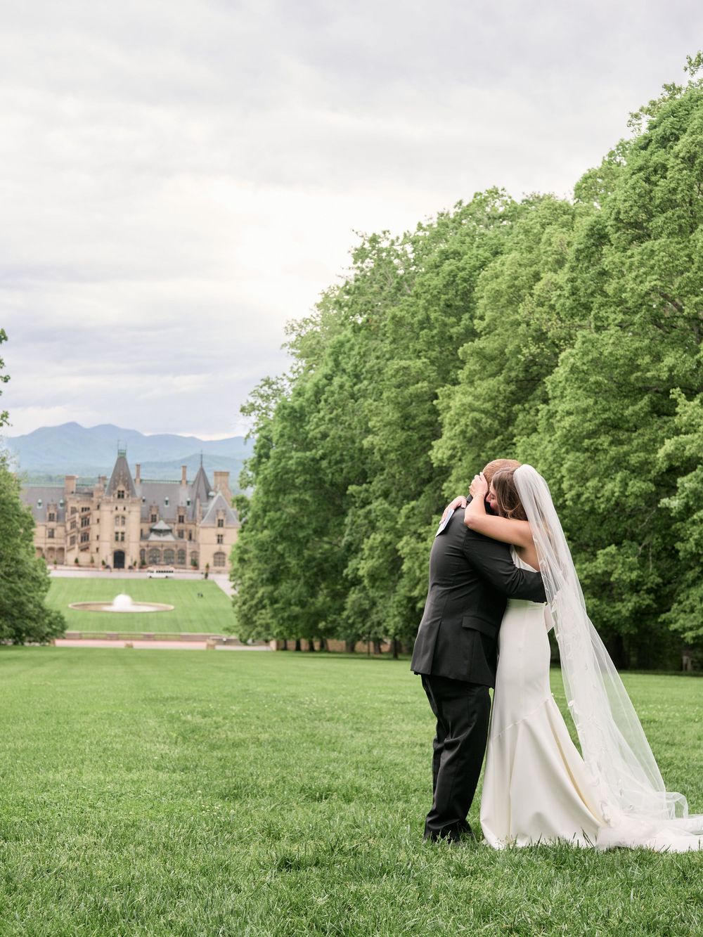 Couple in wedding attire pose together on a vast green lawn with a grand castle-like building in the background.