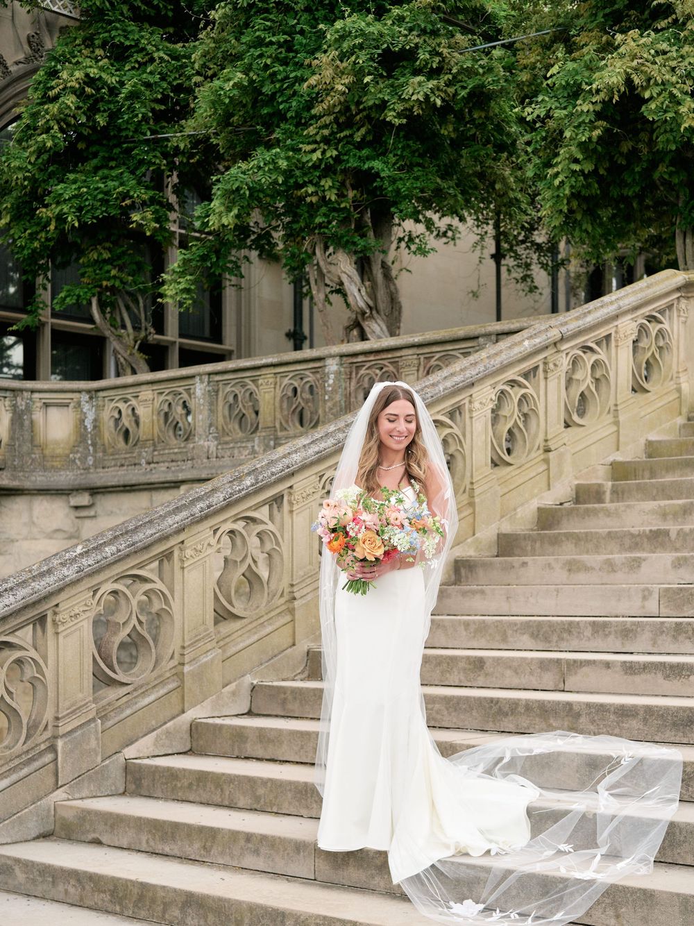 A bride in a flowing white dress and colorful bouquet sits gracefully on elegant stone stairs surrounded by green foliage.
