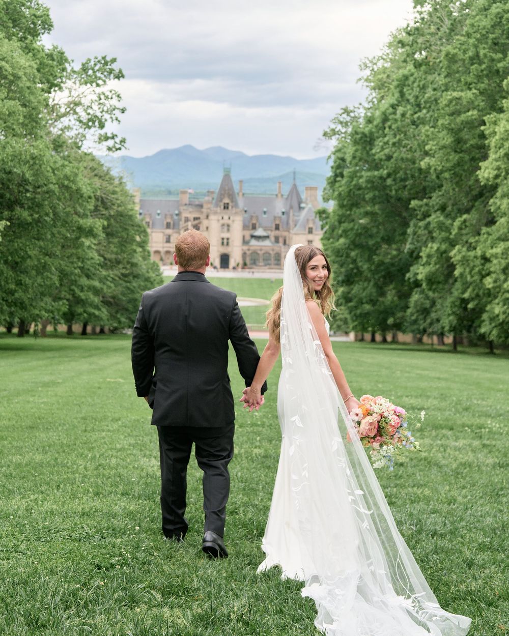 A wedding couple strolling on a manicured lawn with the elegant Biltmore Estate castle visible in the background.