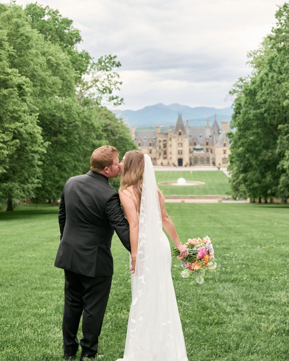 Couple in wedding attire embrace and kiss on expansive green lawn with majestic castle in background at Biltmore Estate.