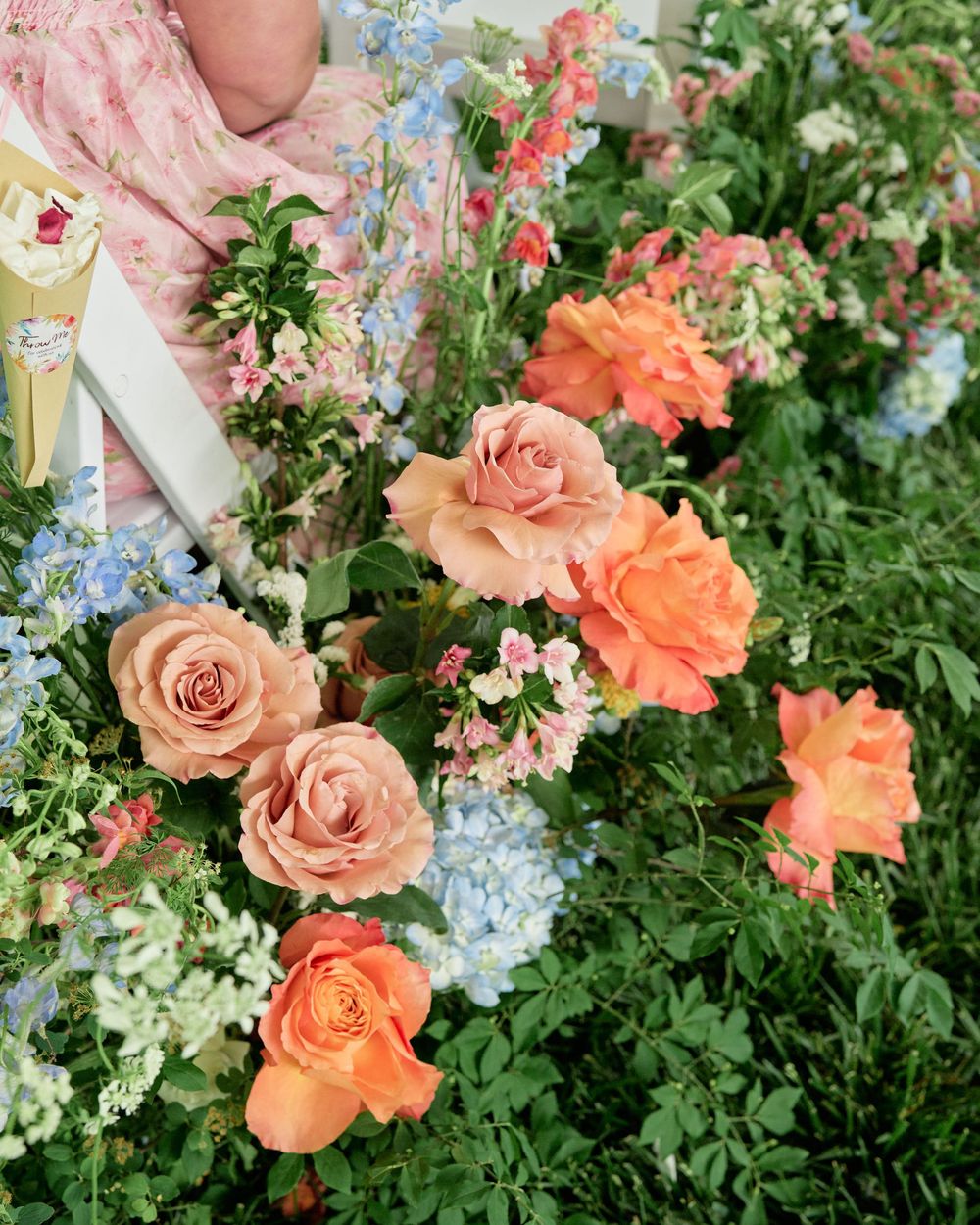 Garden arrangement with peachy-orange roses, delicate blue flowers, and white blooms against lush green foliage.