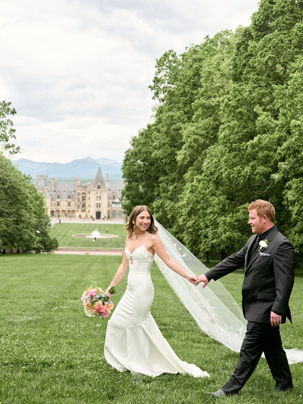 A wedding couple dances and twirls playfully on a green lawn with a stately building visible in the distance.
