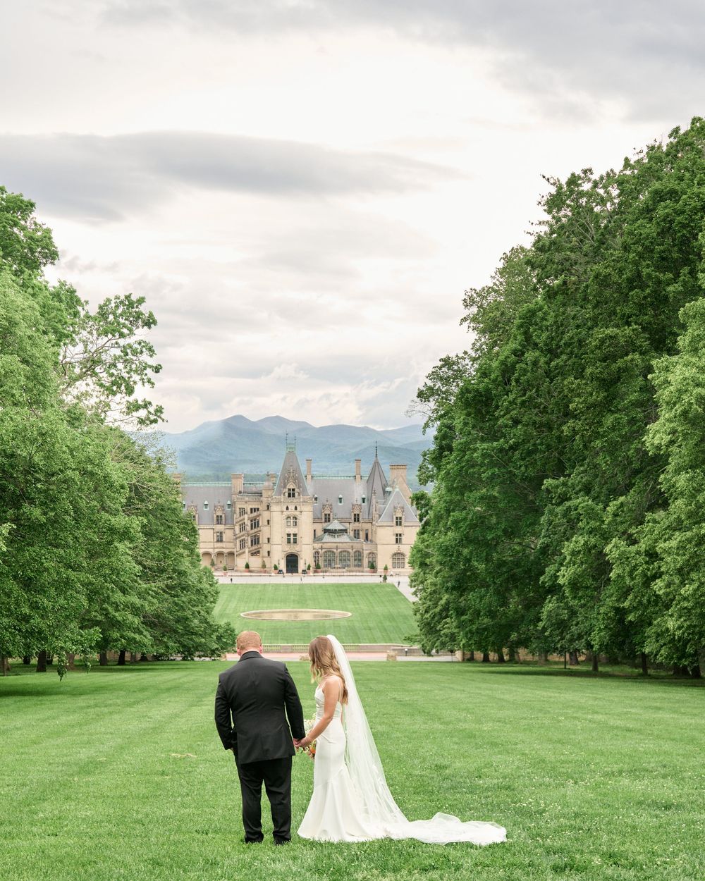 A couple walks across a sprawling green lawn with a historic castle estate visible in the distance under cloudy skies.