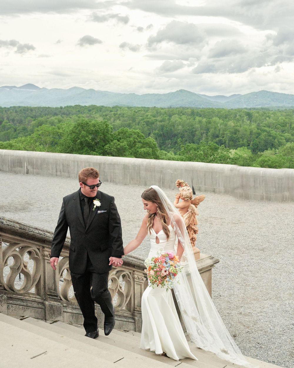 A wedding couple and wedding party in elegant attire pose thoughtfully on a stone terrace overlooking mountain scenery.