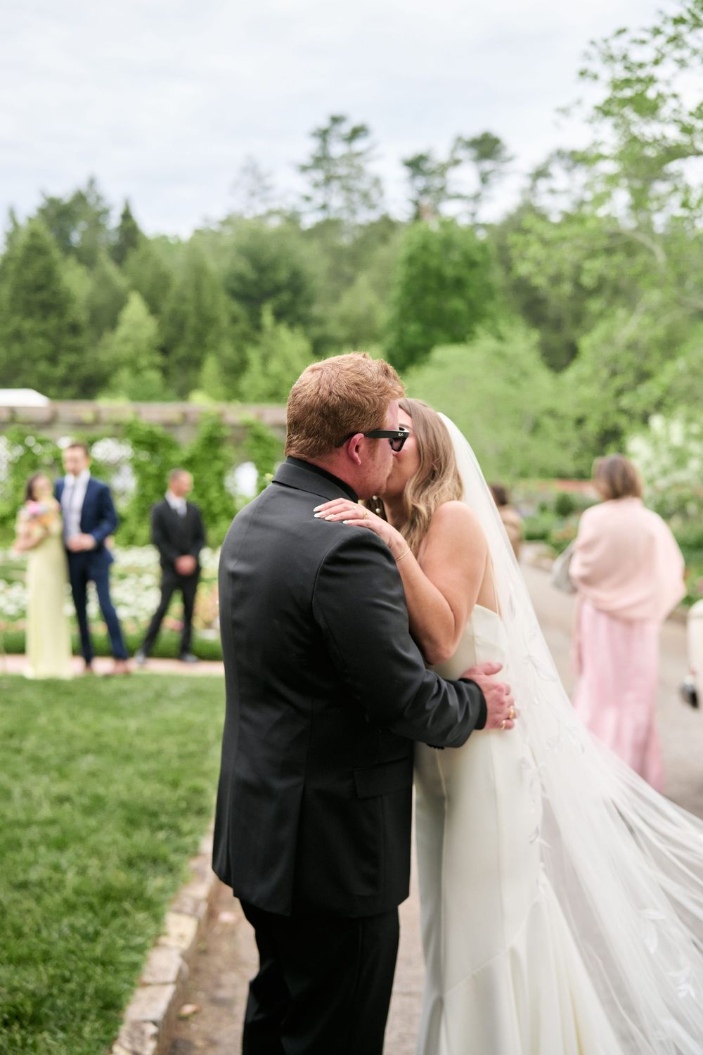 A wedding couple shares an intimate kiss outdoors while surrounded by trees and wedding guests in soft focus background.