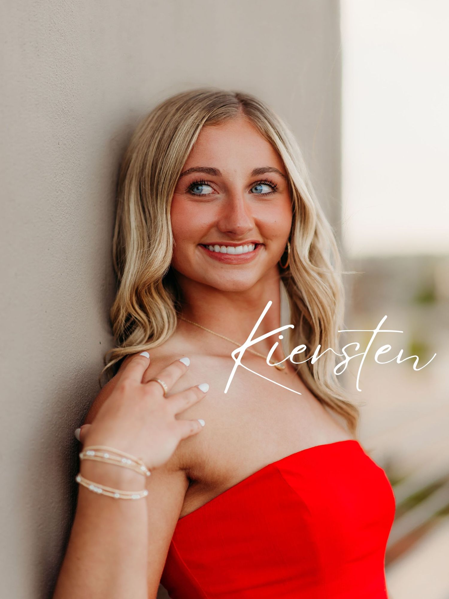 A strapless red evening dress is showcased against a neutral wall with delicate bracelet accessories.