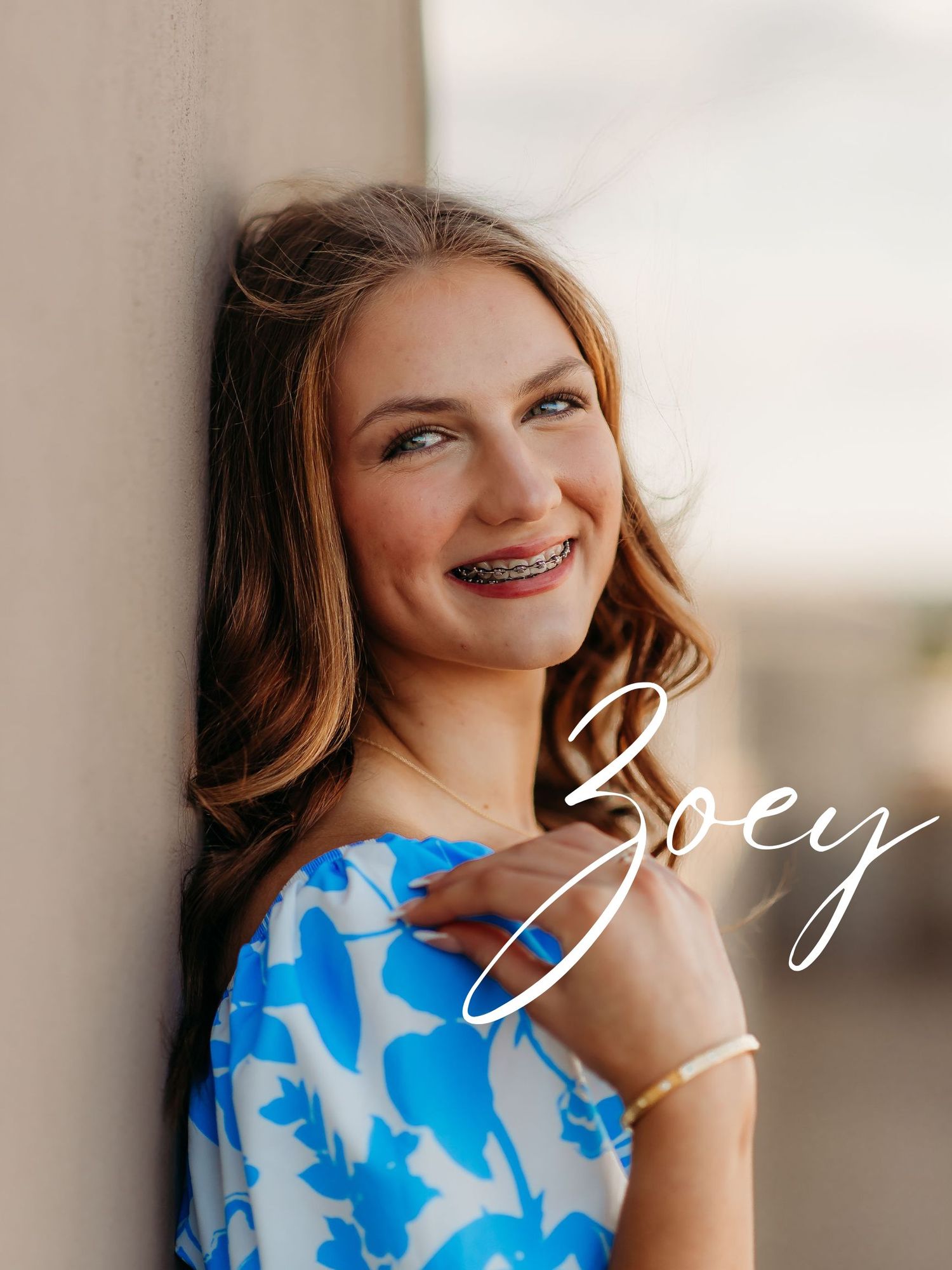 A smiling young person wearing braces and a blue and white patterned dress poses against a light wall.