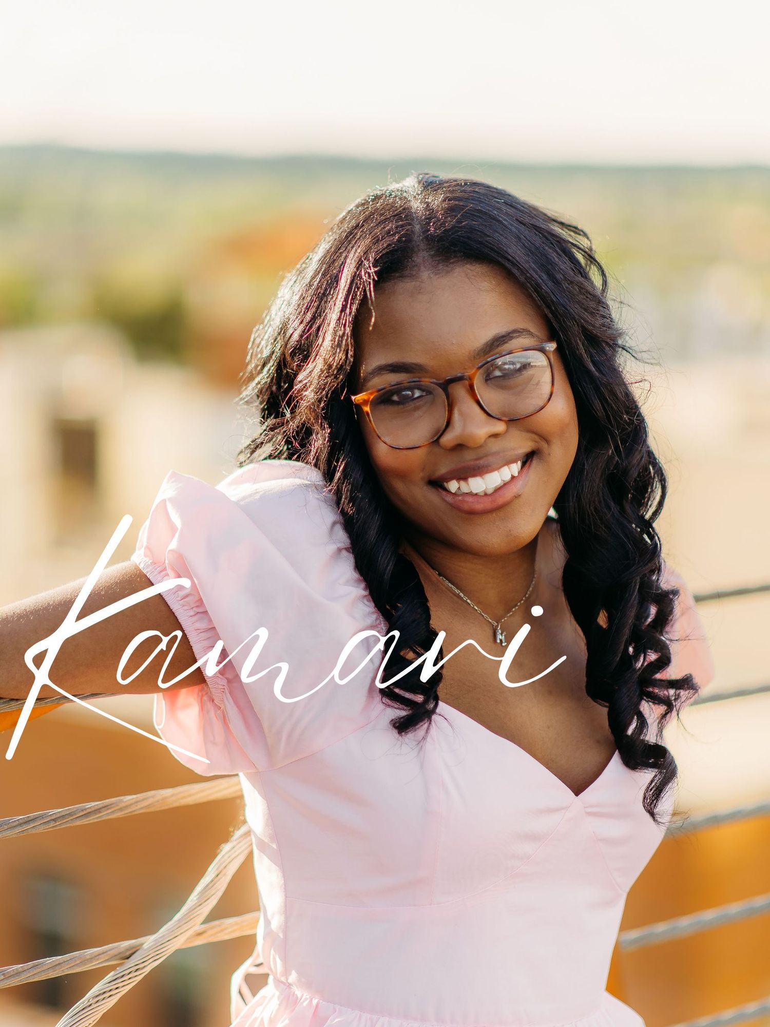 Portrait of someone wearing glasses and a pink top, smiling warmly in natural outdoor lighting.
