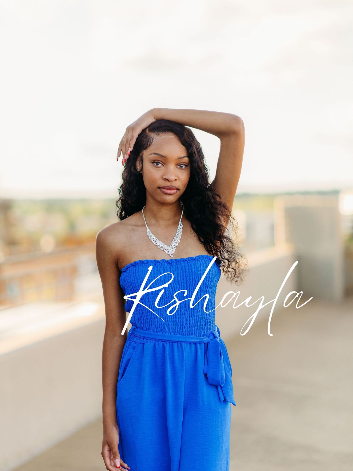 A person wearing a bright blue strapless jumpsuit and silver necklace poses against a rooftop backdrop.
