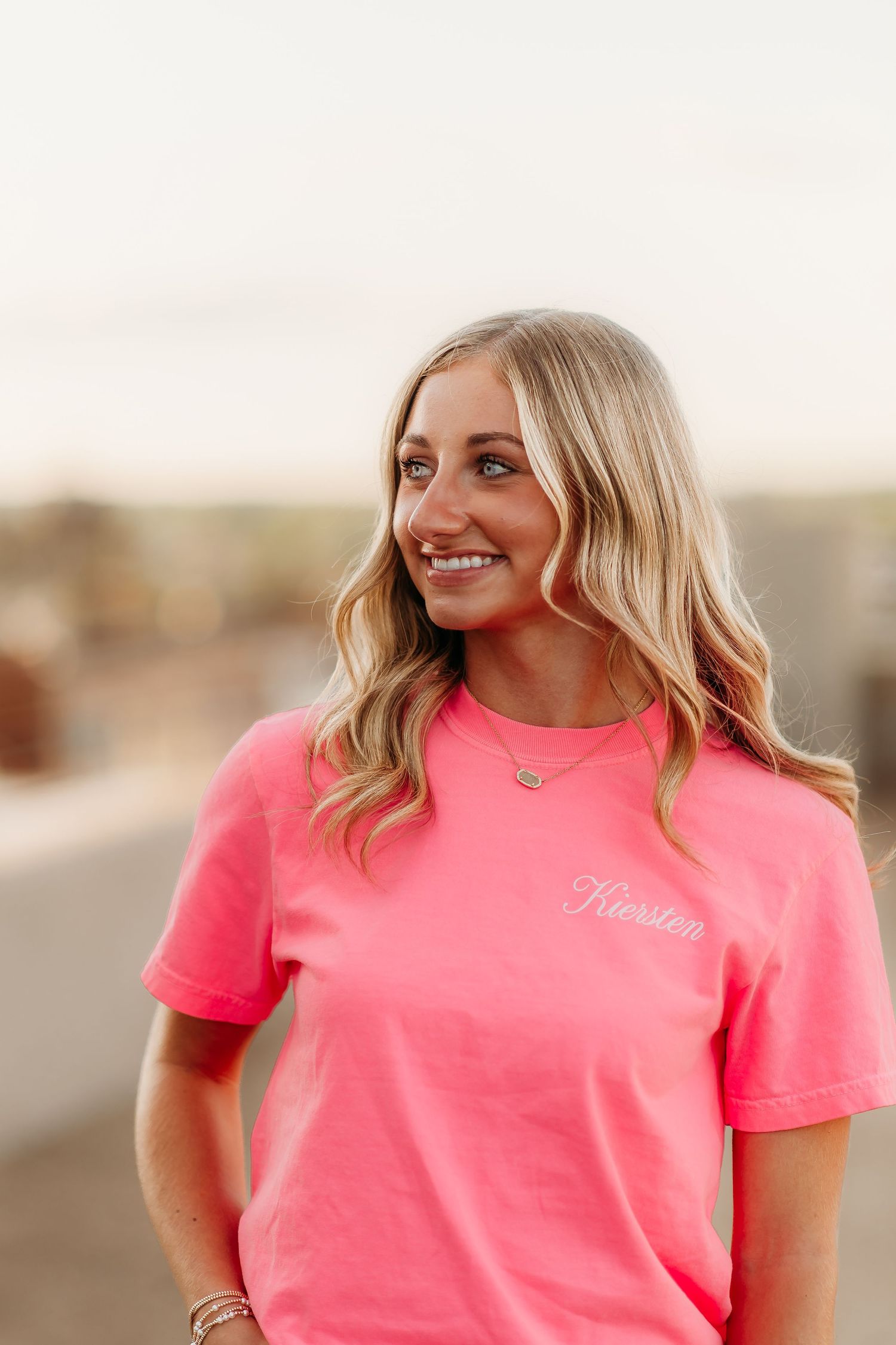 Woman wearing bright coral pink t-shirt smiles outdoors with wavy blonde hair and delicate necklace.